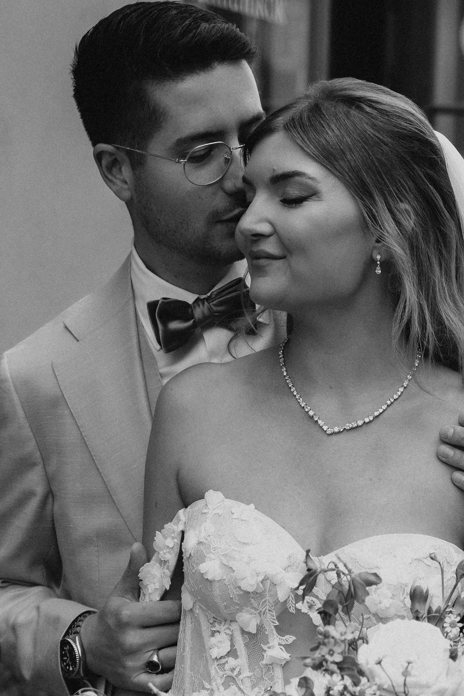 Groom in light suit with bow tie tenderly embracing blonde bride holding floral bouquet in black and white.
