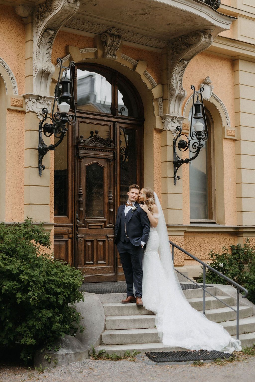 Bride and groom pose on ornate building steps, she in a flowing white gown with veil, he in a dark suit.