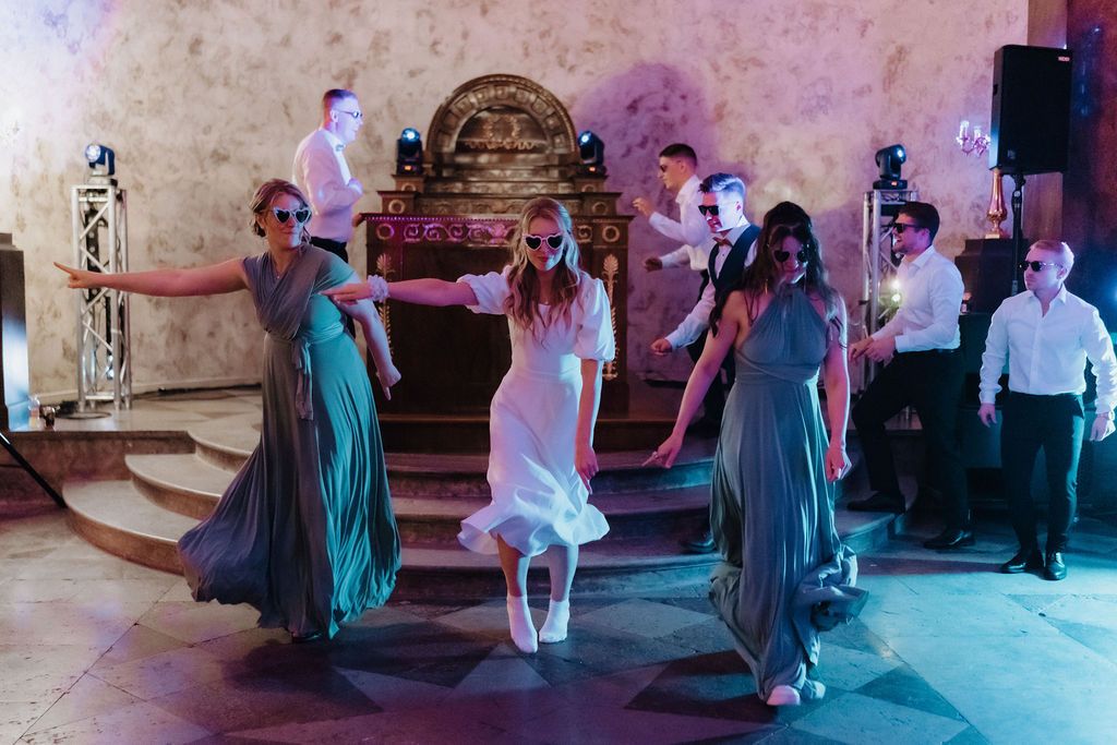 Bridesmaids in grey gowns and bride in white perform synchronized dance routine at wedding reception.