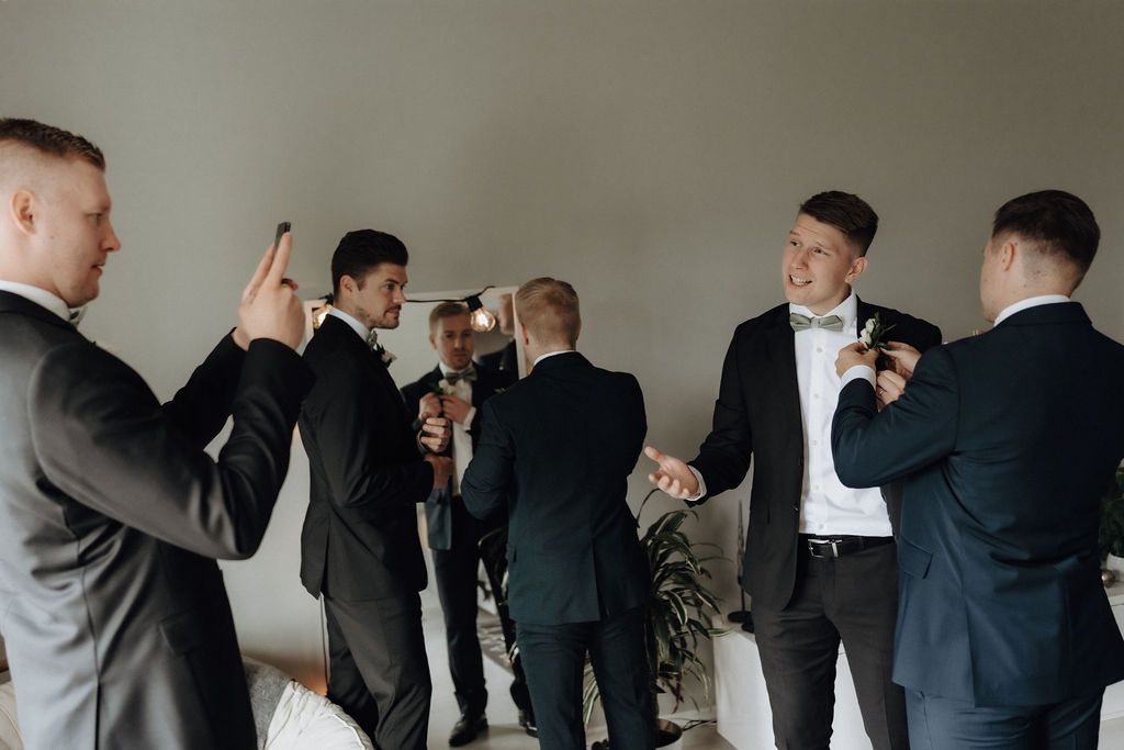 Groomsmen in black suits celebrate together before a wedding ceremony in a bright room.