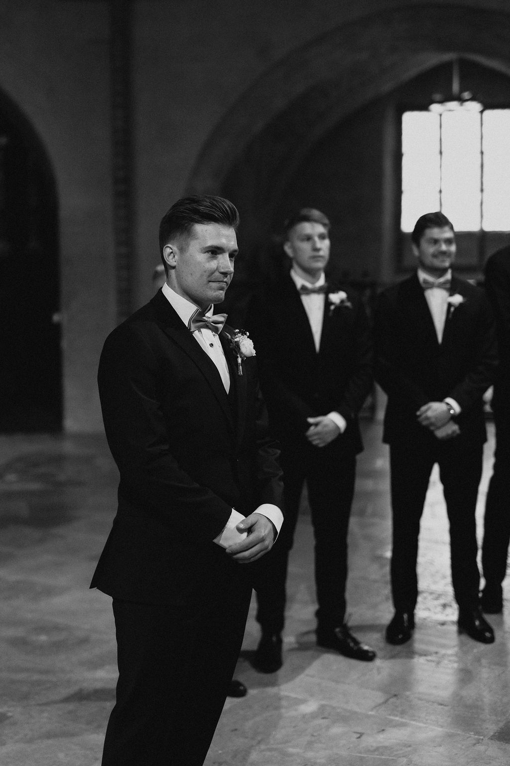 Groom in black tuxedo with boutonniere waits anxiously at altar with groomsmen behind him.