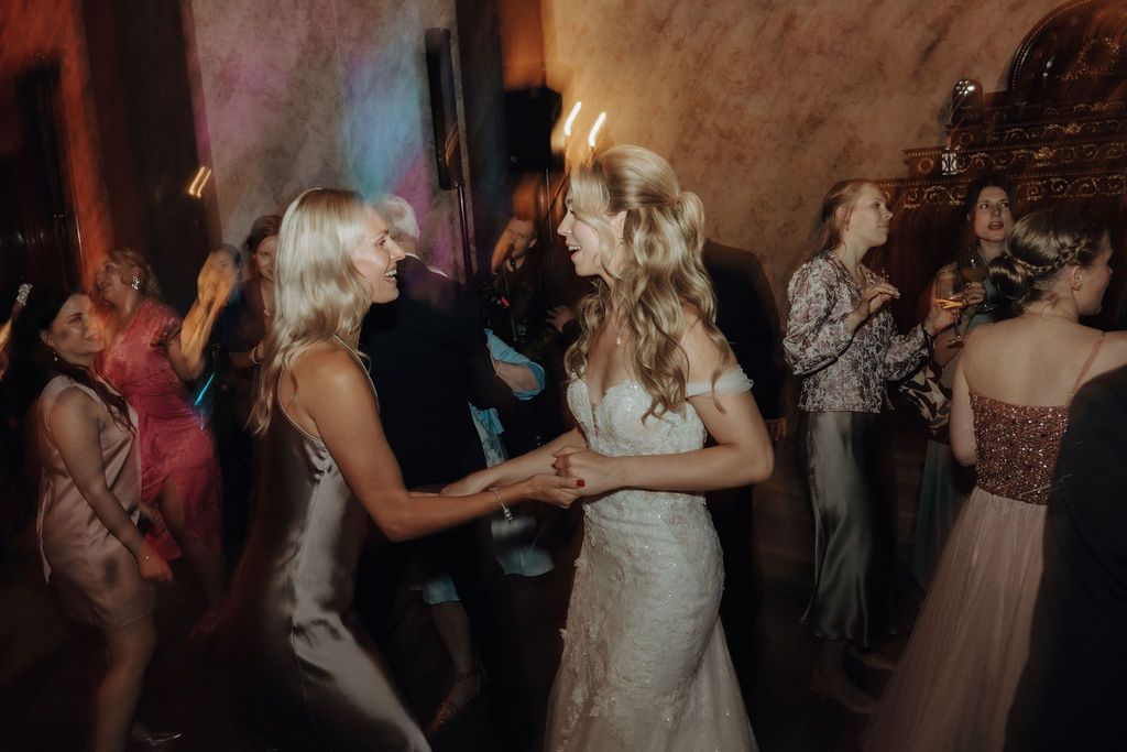 Bride in lace gown dances joyfully with blonde guest at wedding reception.