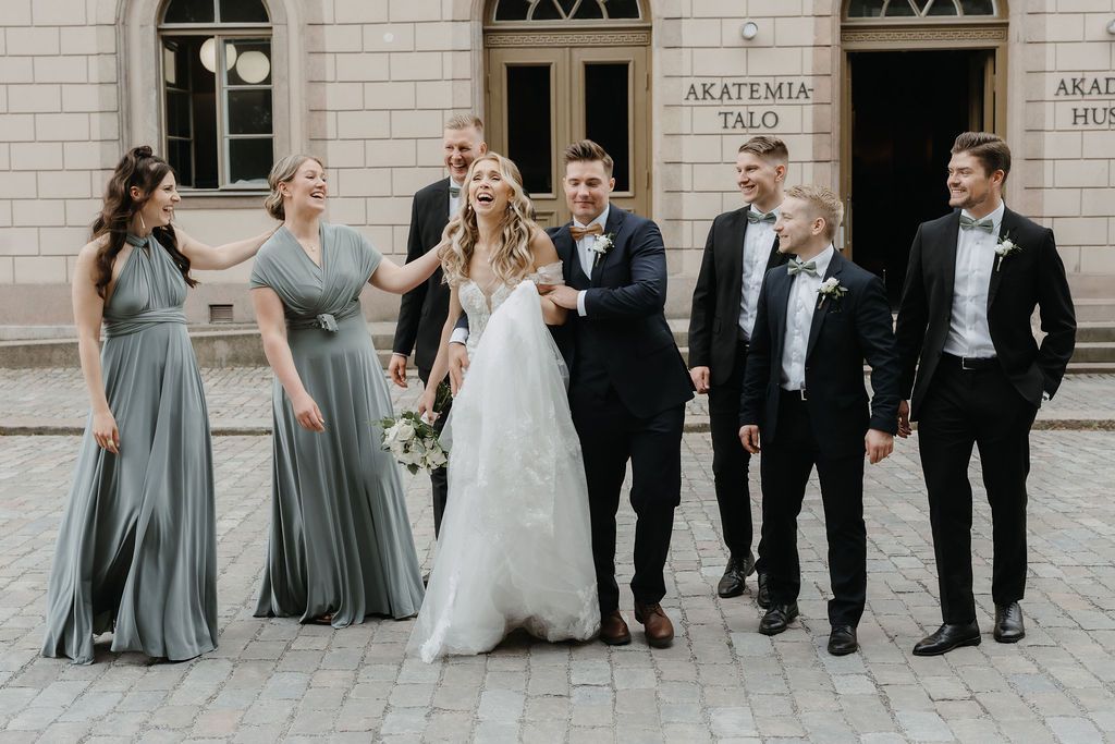 Joyful wedding party poses outside Akatemia Talo with bride in lace gown and bridesmaids in sage dresses.