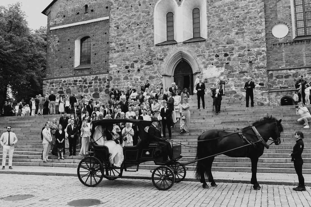 Black and white photo of wedding party with horse-drawn carriage outside historic stone church.
