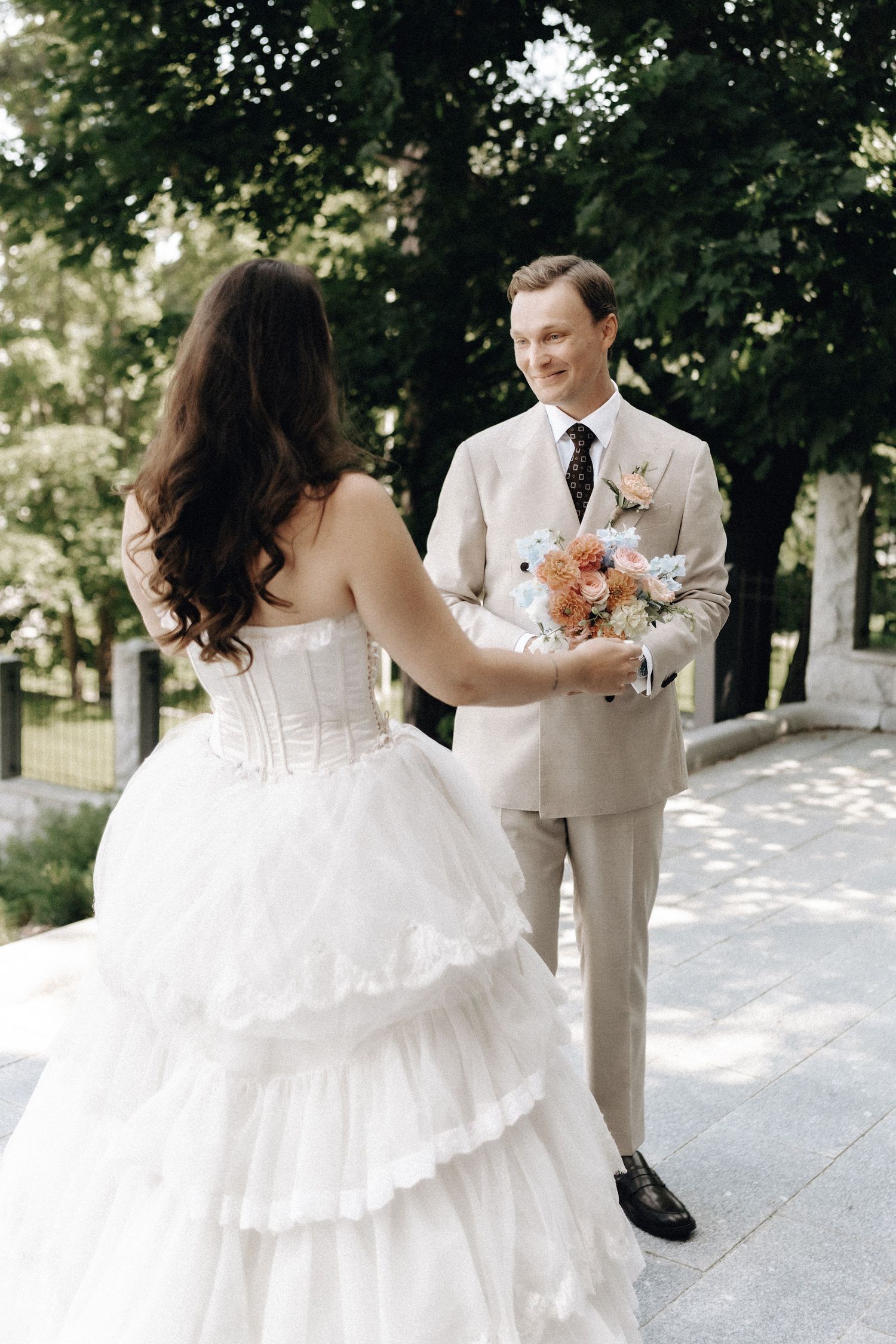 Smiling groom in beige suit holding pastel bouquet facing bride in strapless layered white gown outdoors.