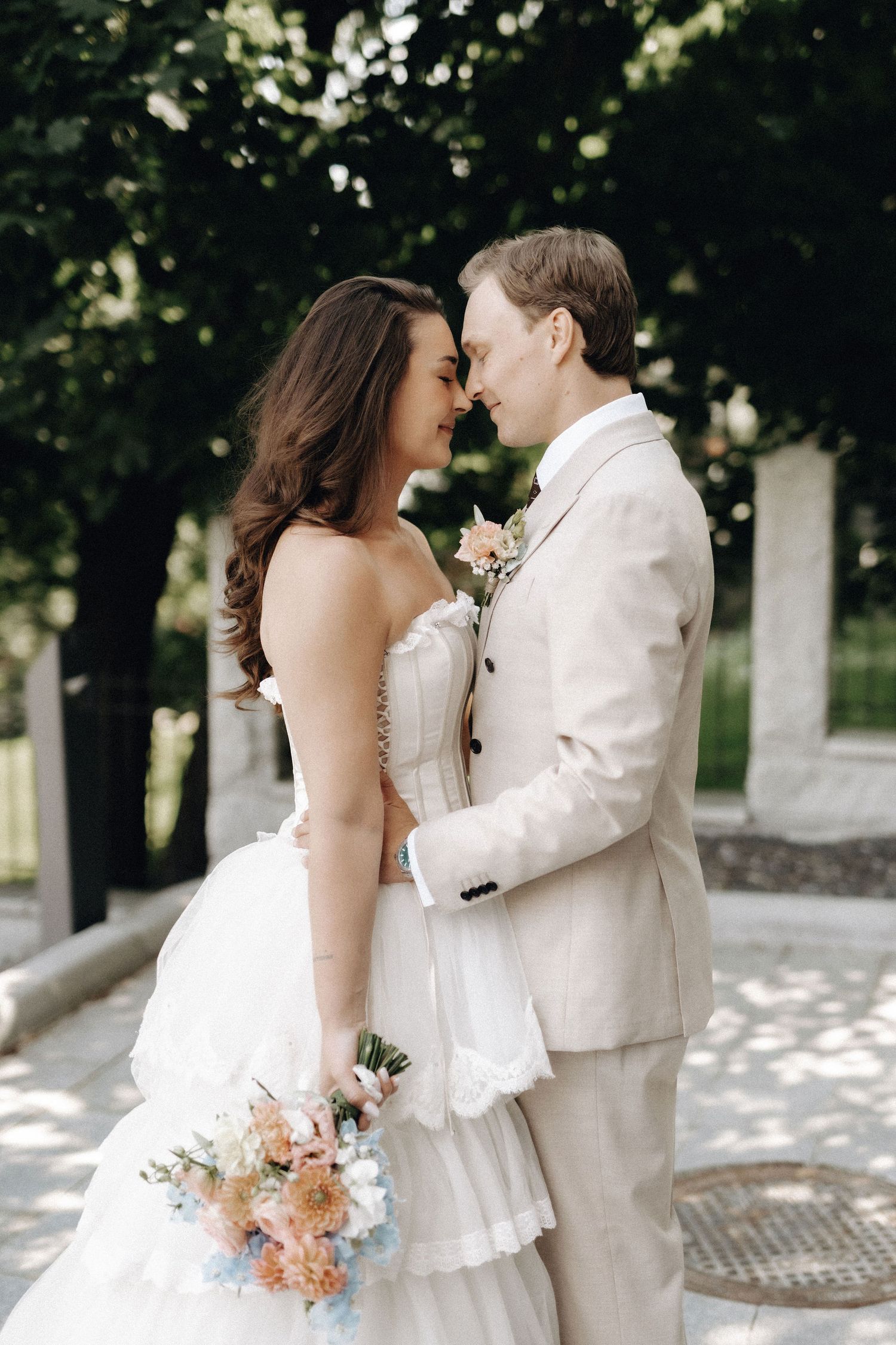 Bride and groom touching noses romantically outdoors, she holds a pastel bouquet while he wears a beige suit.