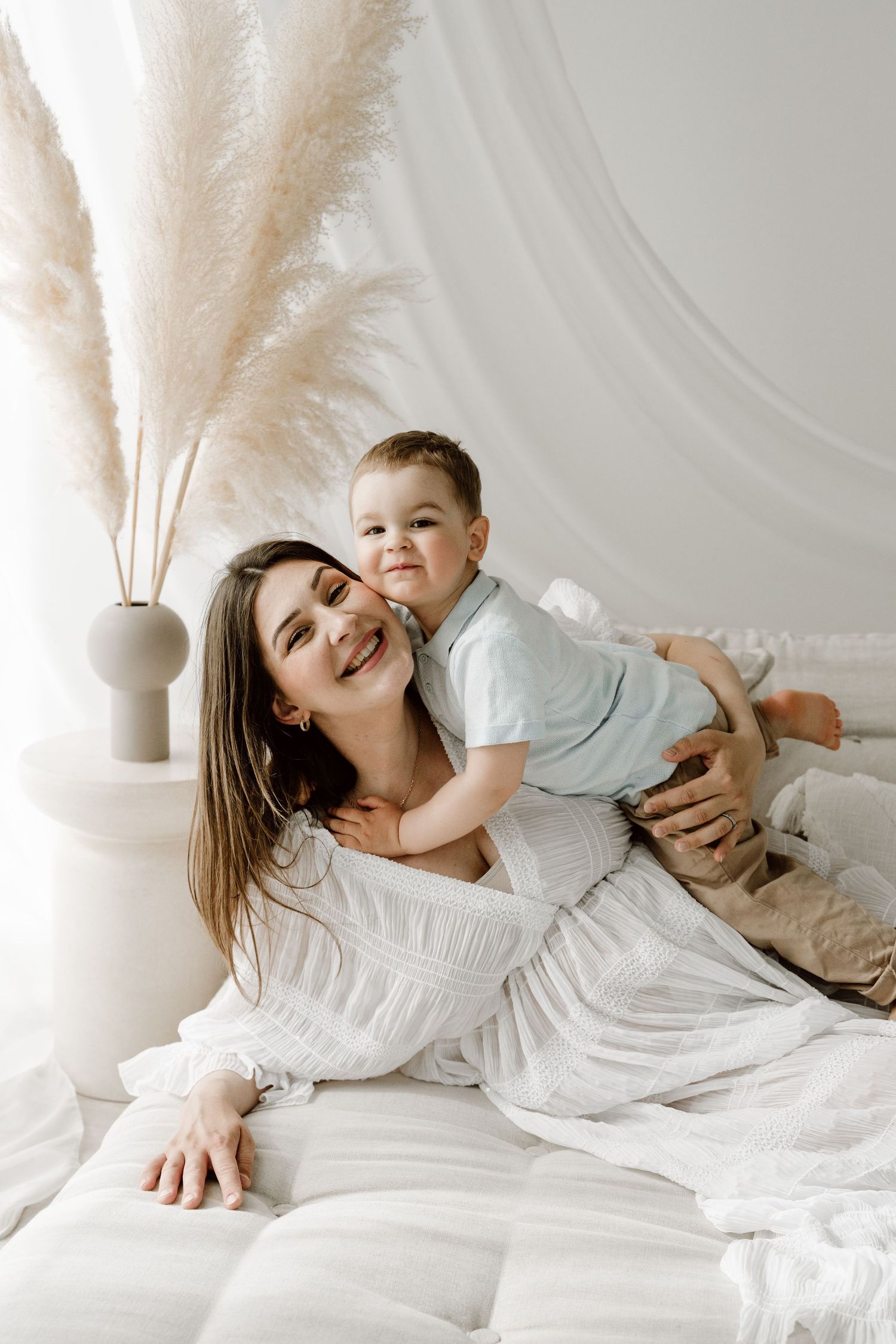 A happy mother in a white dress plays with her toddler son on a bed with pampas grass decor.