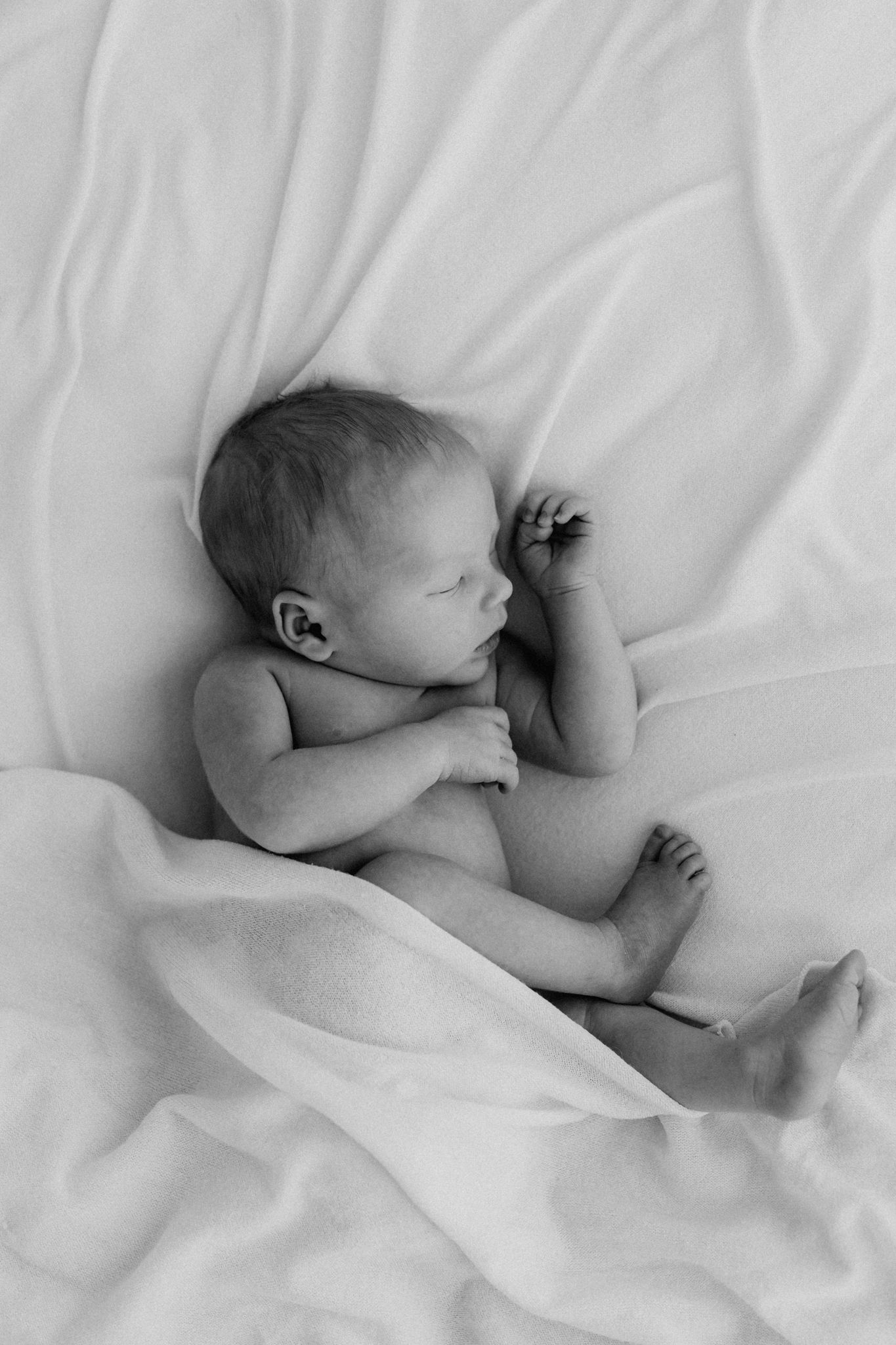 Black and white photo of a peaceful naked newborn baby sleeping on soft white fabric.
