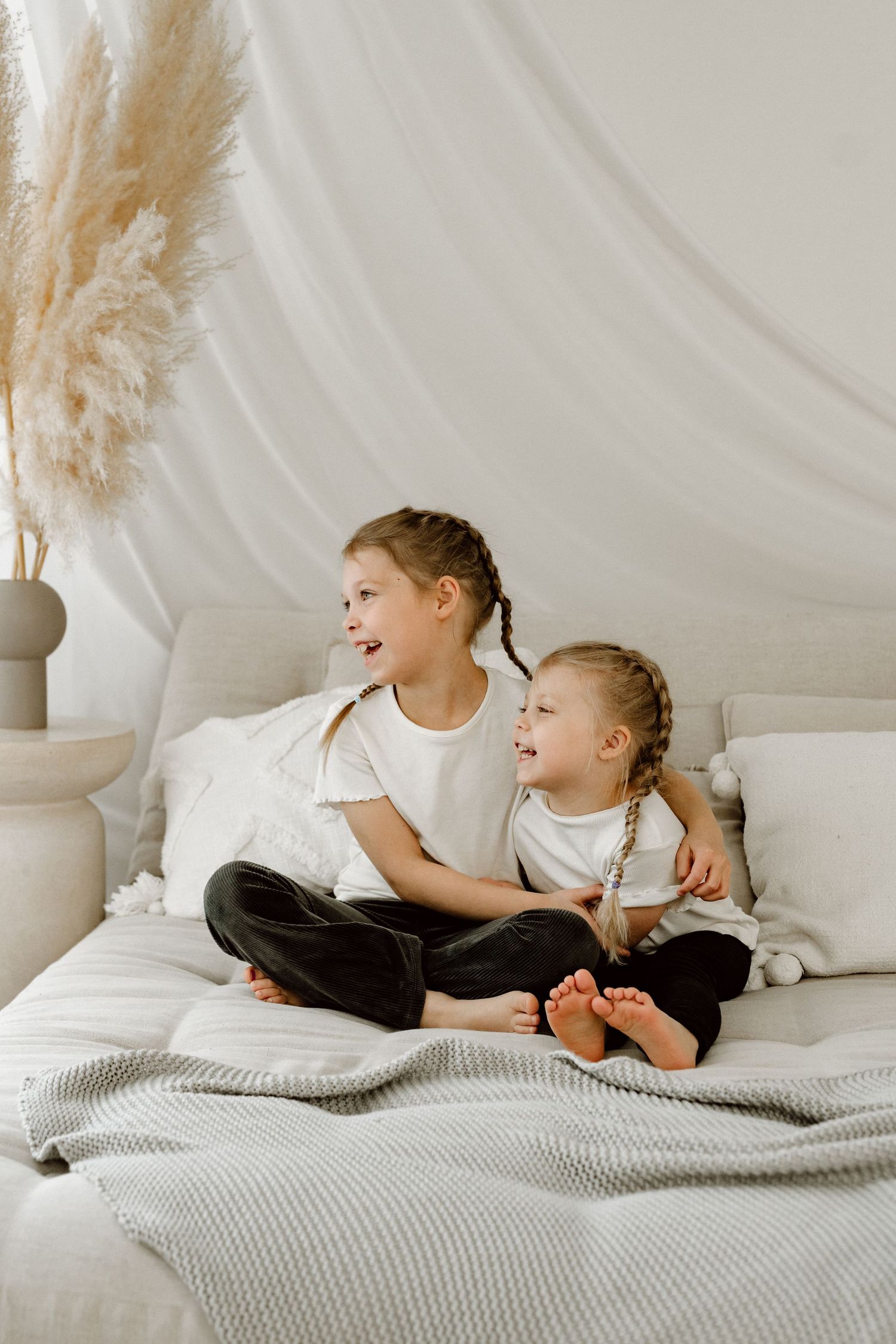 Two young sisters with braids sitting together on white quilted bed near pampas grass.