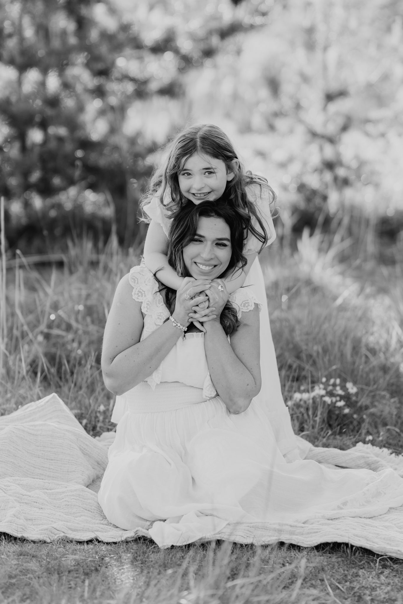 Black and white photo of a girl hugging her mother from behind while sitting on a blanket outdoors.