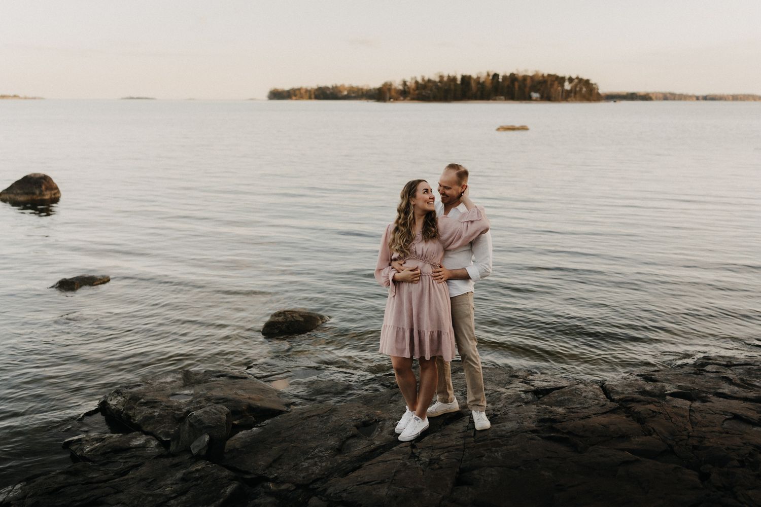 Romantic couple holding each other on rocky lakeside shore with island and soft sunset sky behind them.