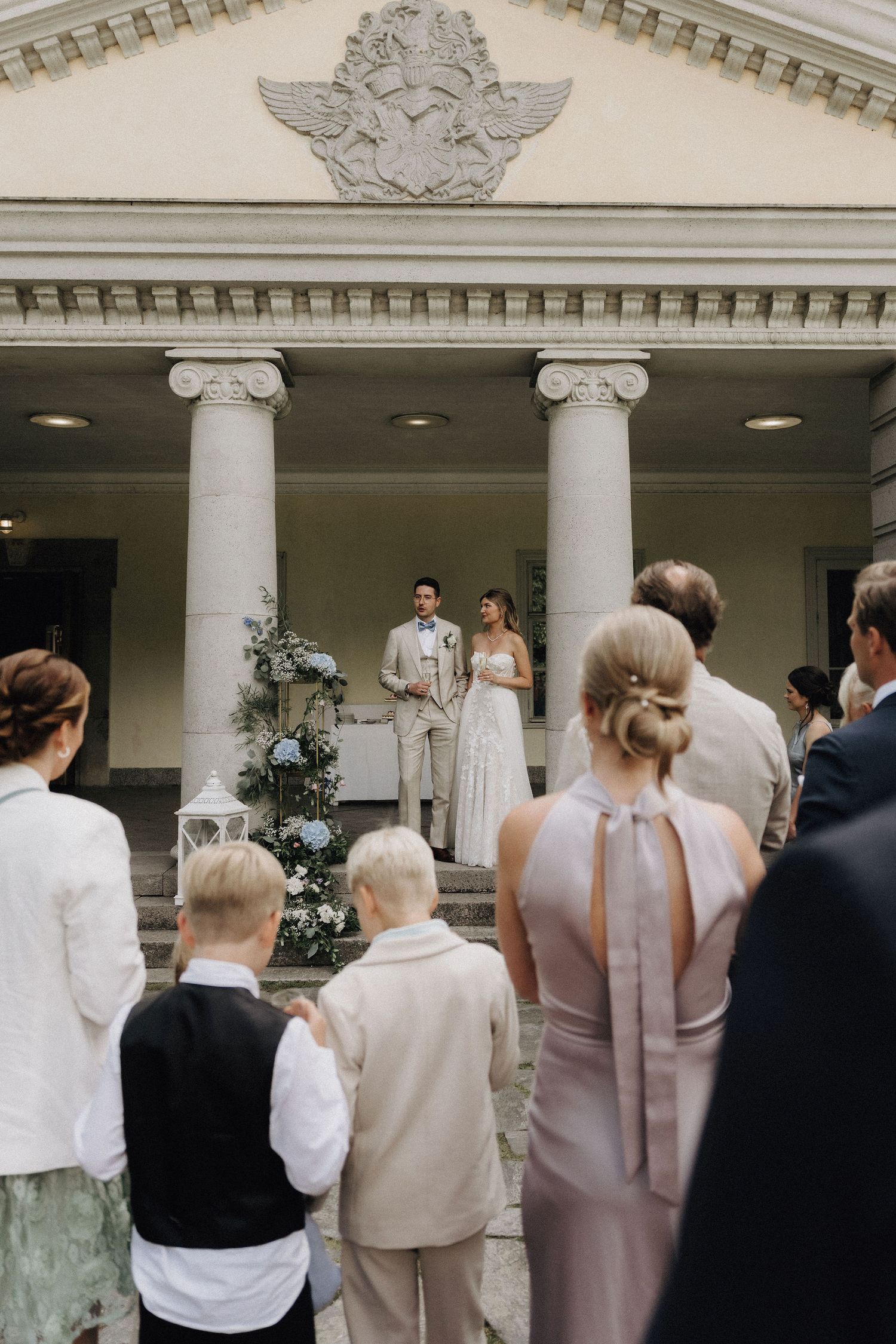 Wedding ceremony outdoors at a neoclassical building with columns as guests watch the couple on steps.
