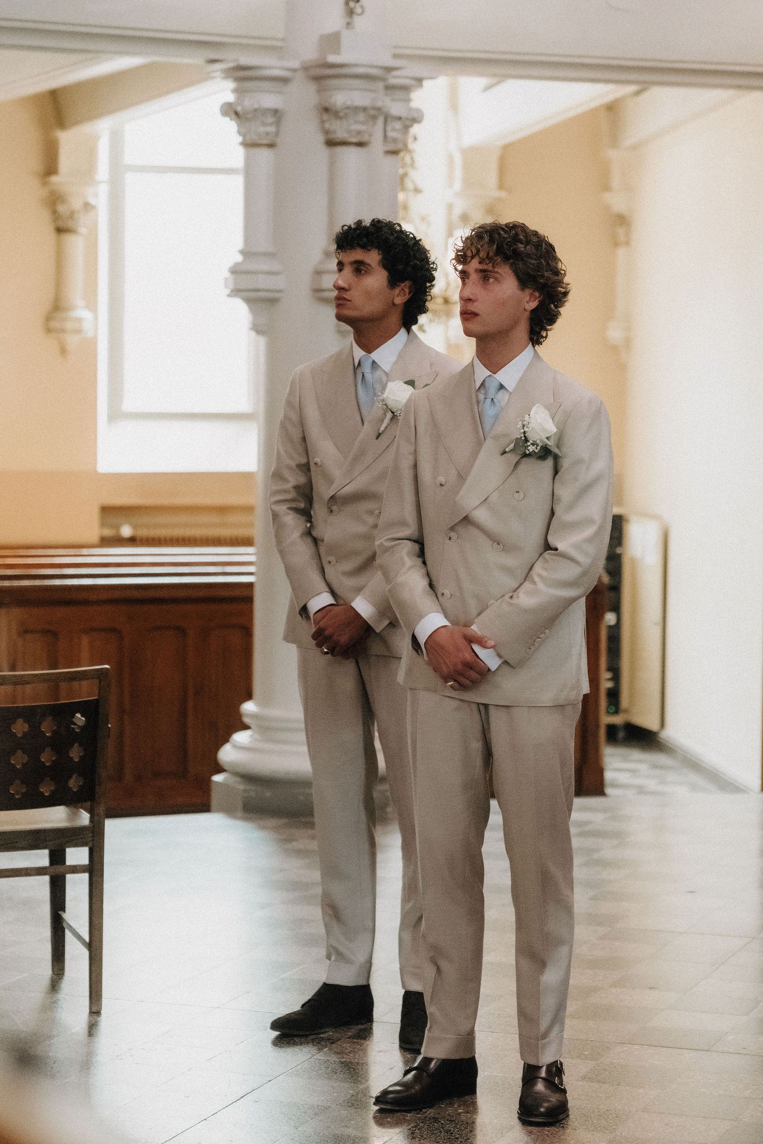 Two groomsmen in matching tan double-breasted suits with white boutonnieres standing in church.