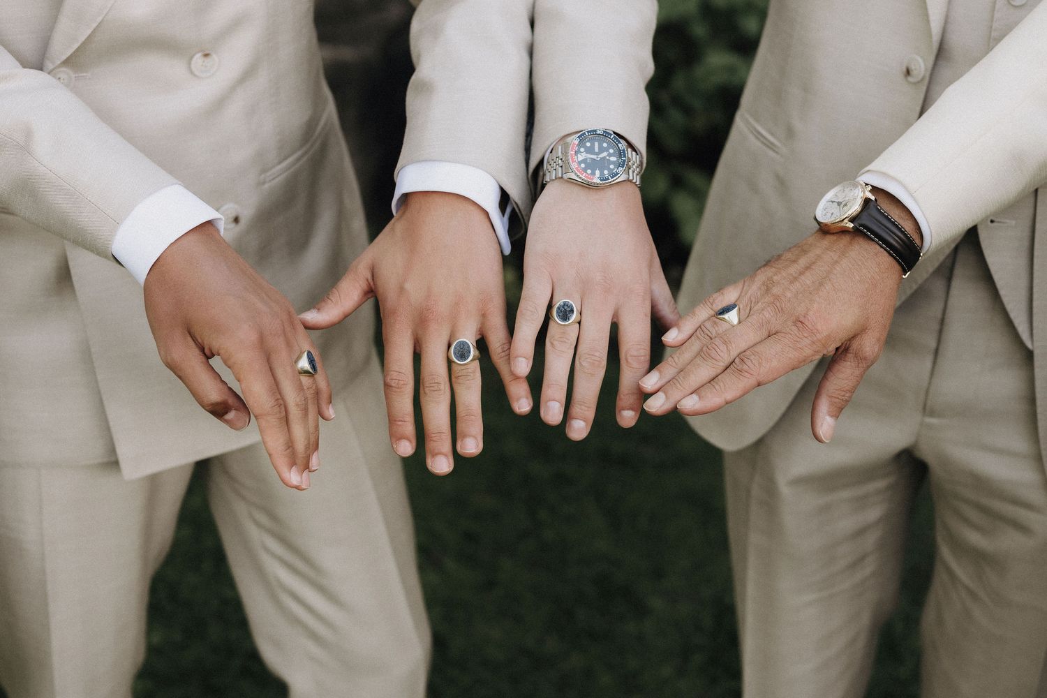 Close-up of three groomsmen showing off rings and watches while wearing beige wedding suits.