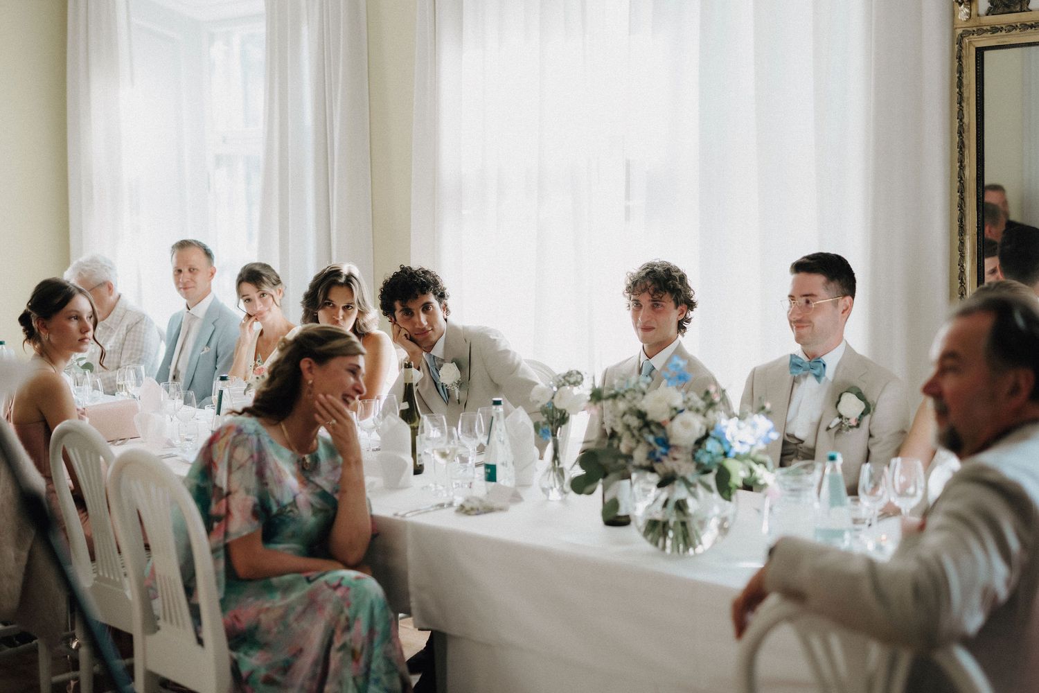 Wedding guests seated at elegantly decorated reception table with floral arrangements in bright room.
