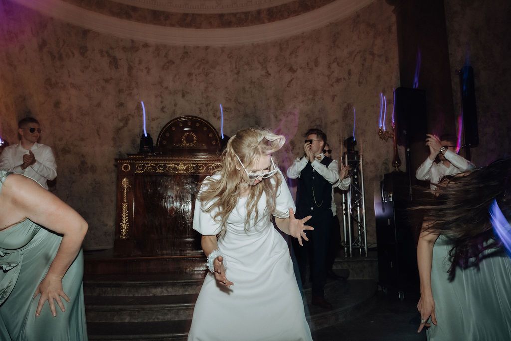 Bride in white dress dancing energetically with guests at a wedding reception in ornate venue.