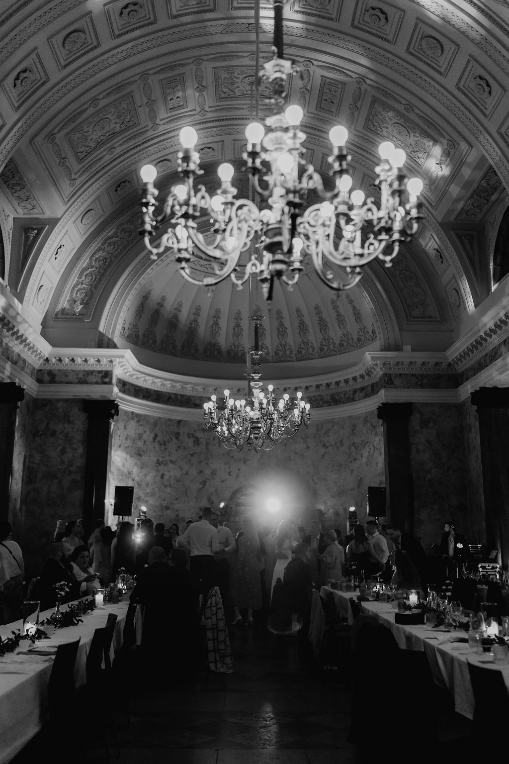 Black and white photo of elegant wedding reception hall with ornate chandeliers and guests dining.