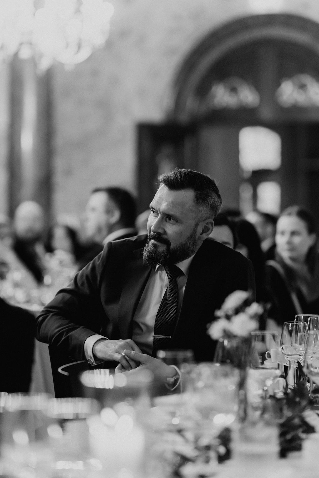 Black and white portrait of a bearded wedding guest in a suit seated at the reception table.