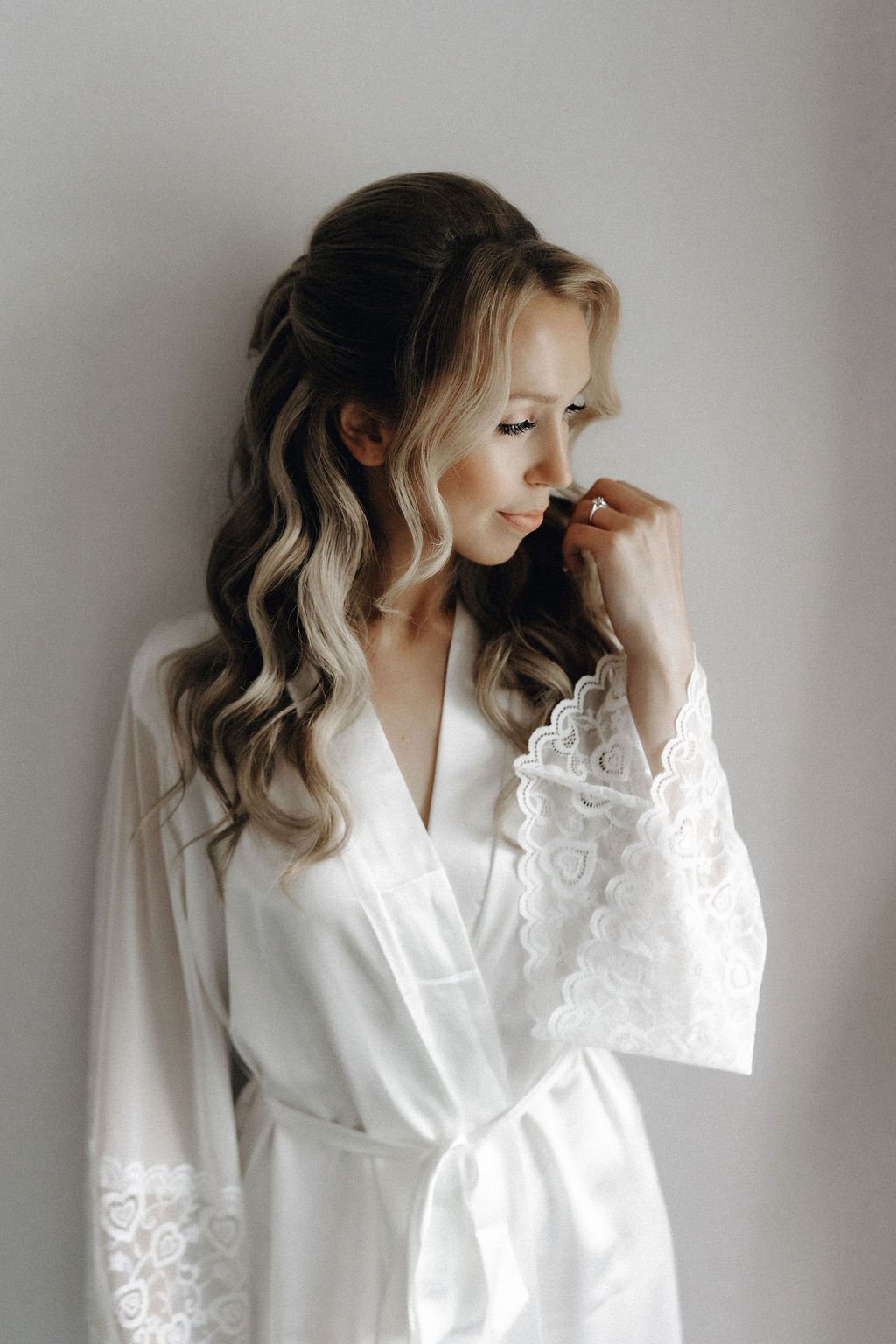 Beautiful blonde bride in white lace robe with elegant half-up curled hairstyle before wedding.