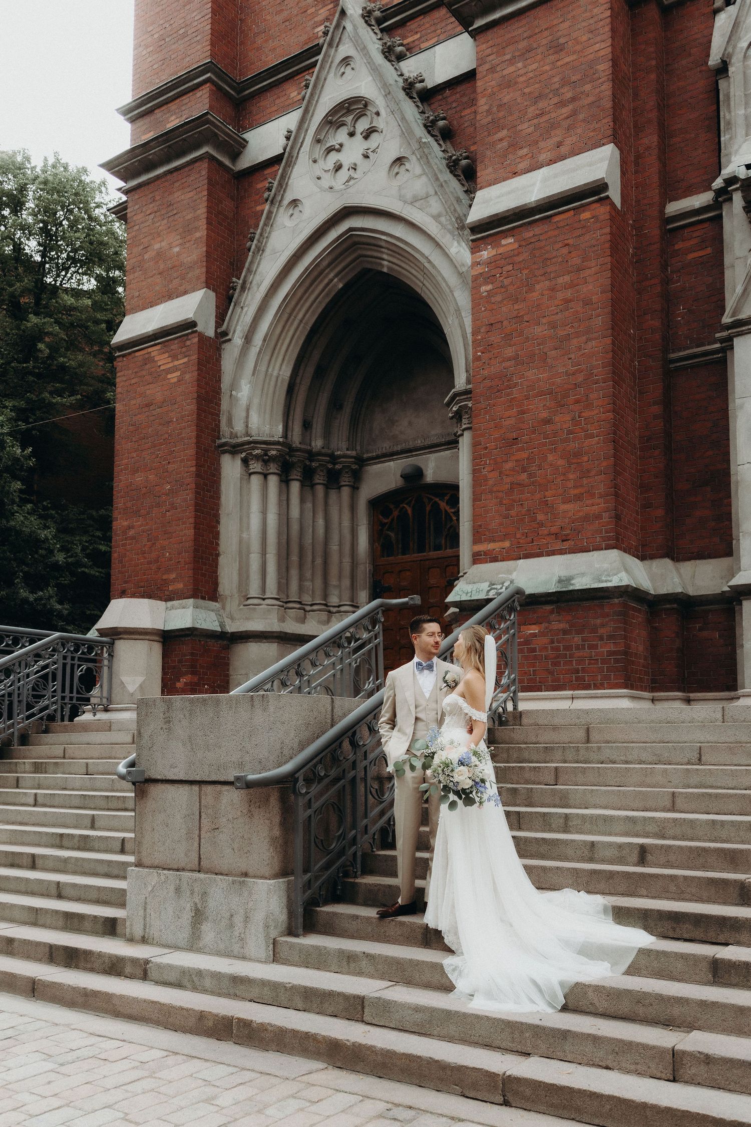 Bride and groom pose on stone steps of a grand red brick Gothic church on their wedding day.