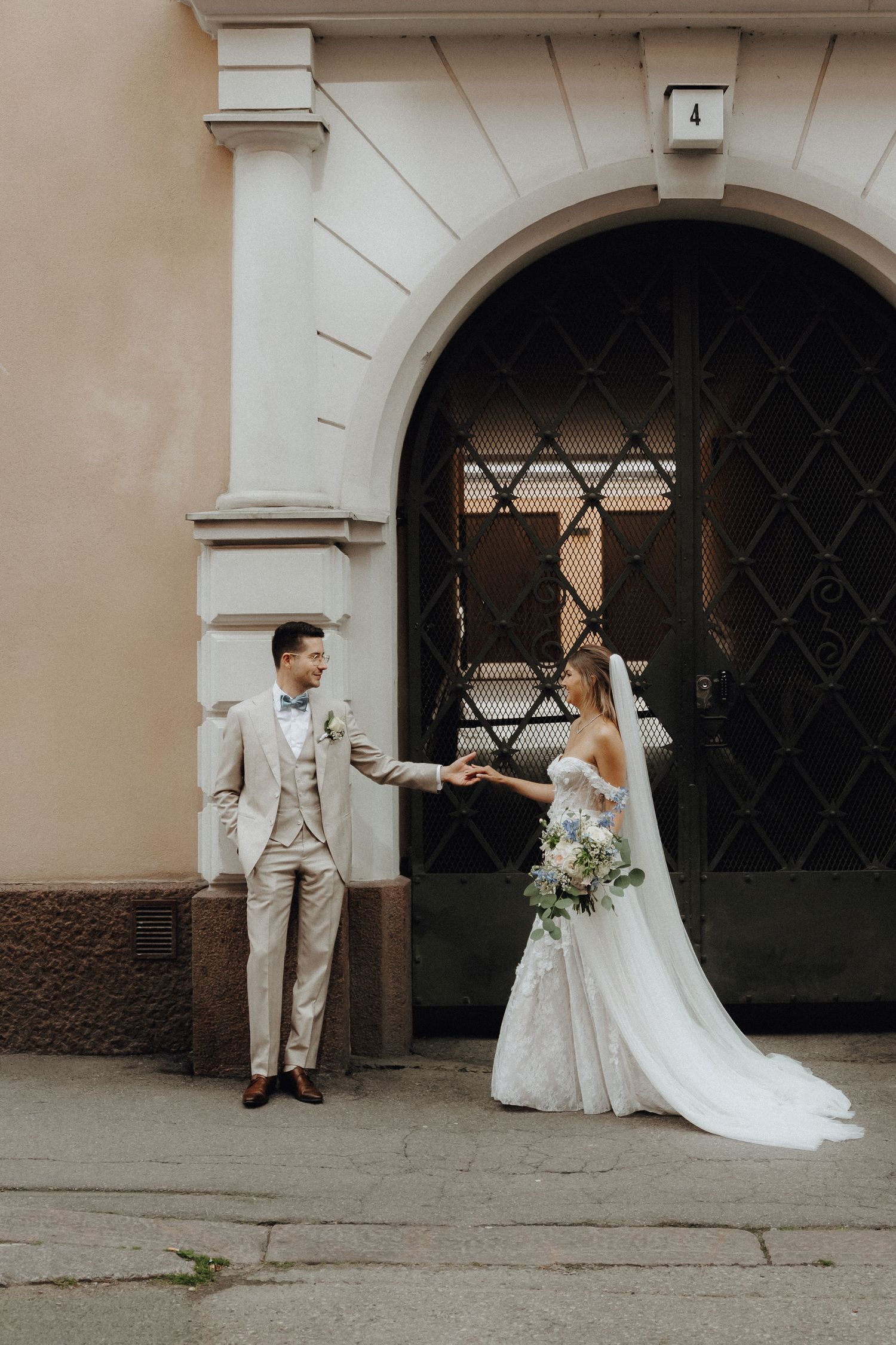 Bride and groom holding hands before an ornate arched doorway on a European city street.