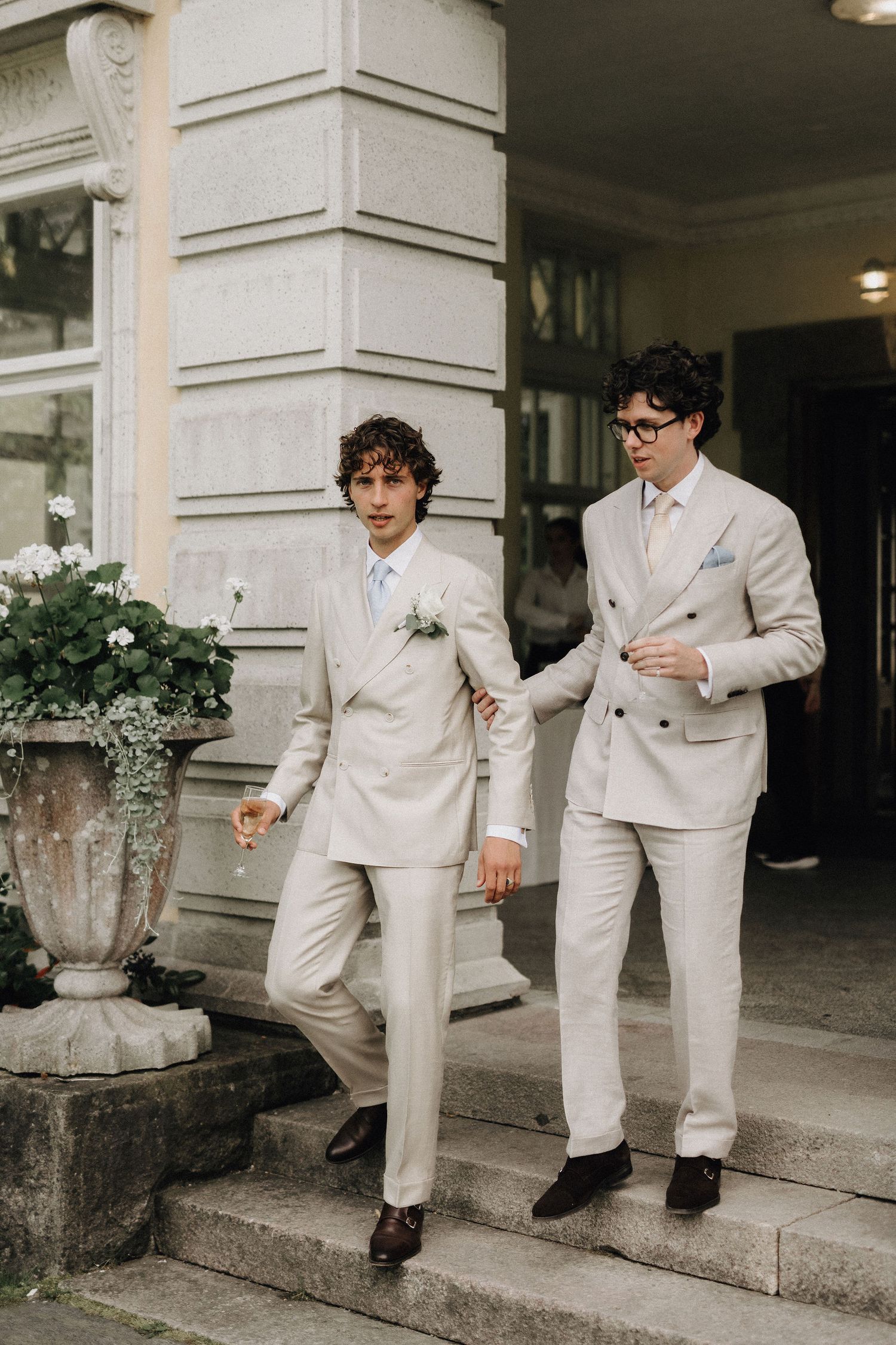 Two grooms in matching cream suits descend stone steps outside a historic building on their wedding day.