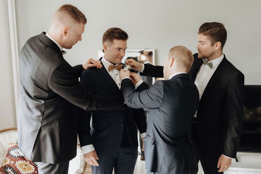 Three groomsmen in black suits helping groom adjust bow tie before wedding ceremony.
