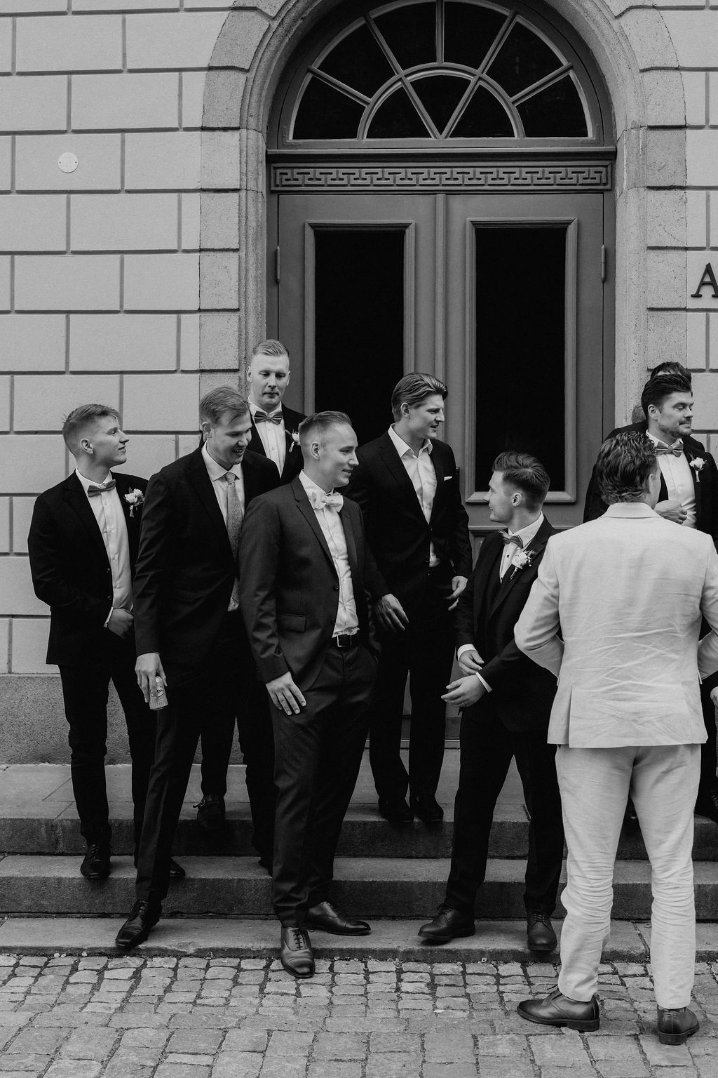 Groom and groomsmen in black tuxedos gather on steps outside a classic stone building.