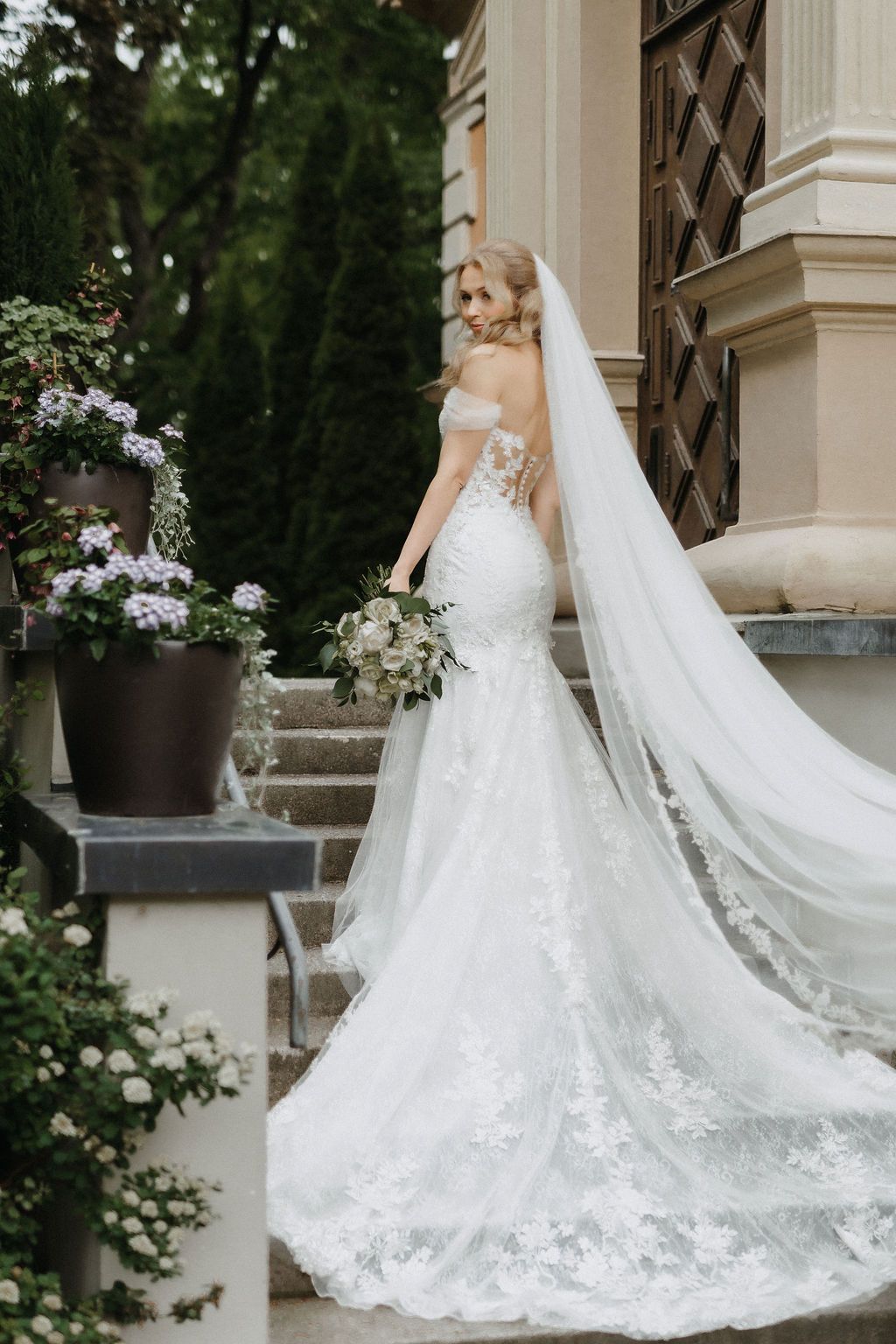 Blonde bride in off-shoulder lace mermaid dress with cathedral veil poses on steps of elegant building.