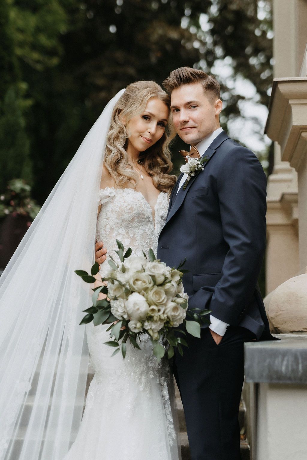 Bride in lace mermaid gown with cathedral veil leans on groom in navy suit holding white bouquet.