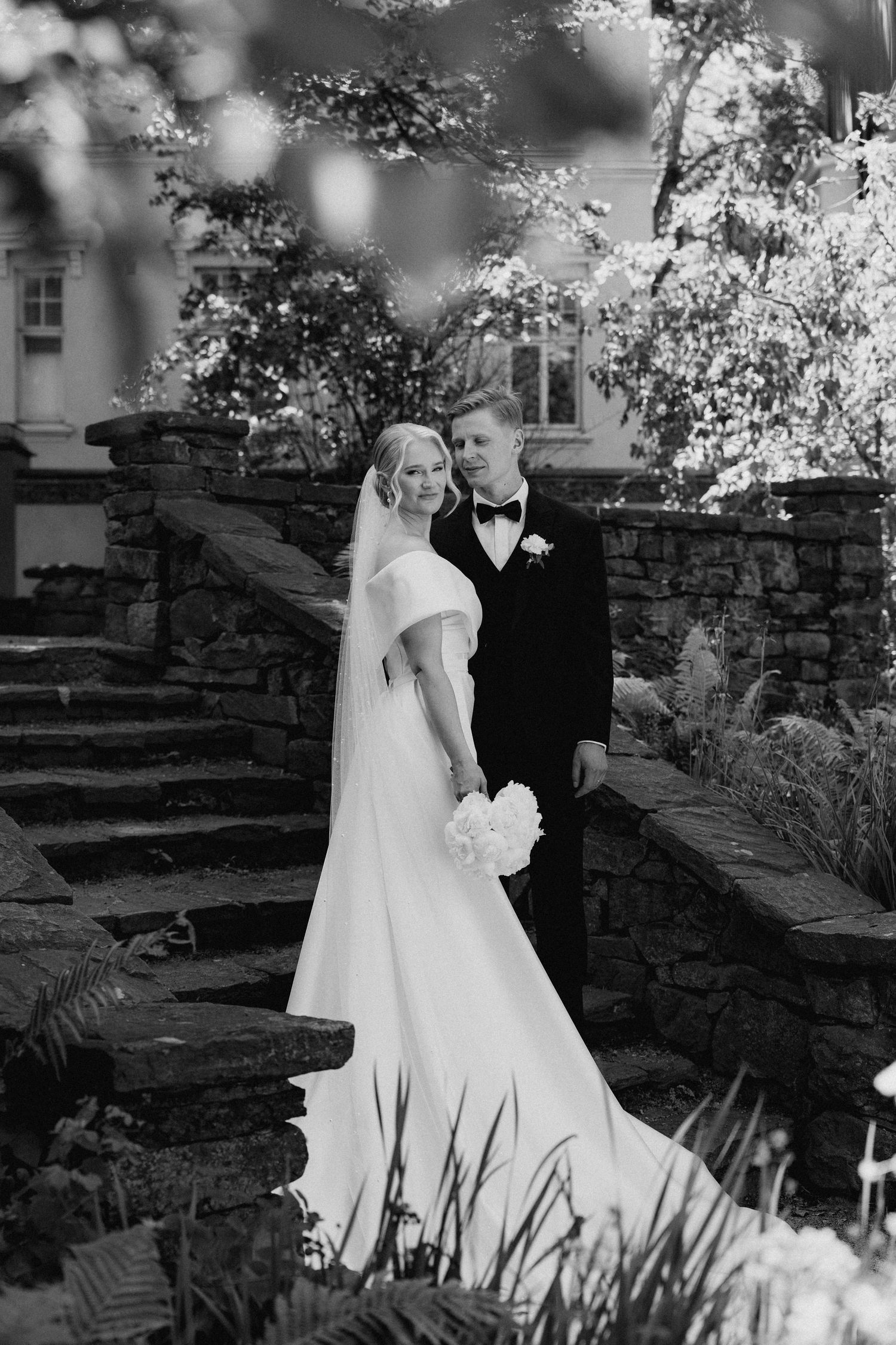 Black and white portrait of elegant bride in off-shoulder gown and groom in tuxedo on stone garden steps.