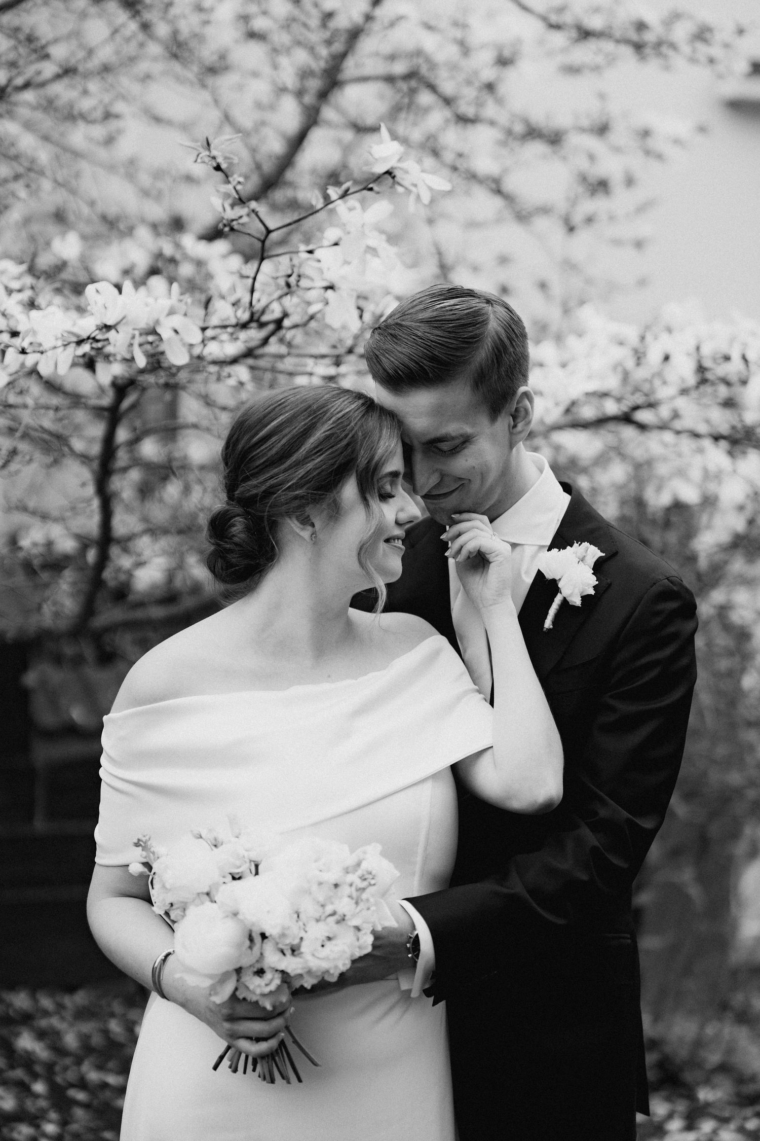 Bride with bouquet and groom share a tender moment under blooming magnolia trees in black and white.
