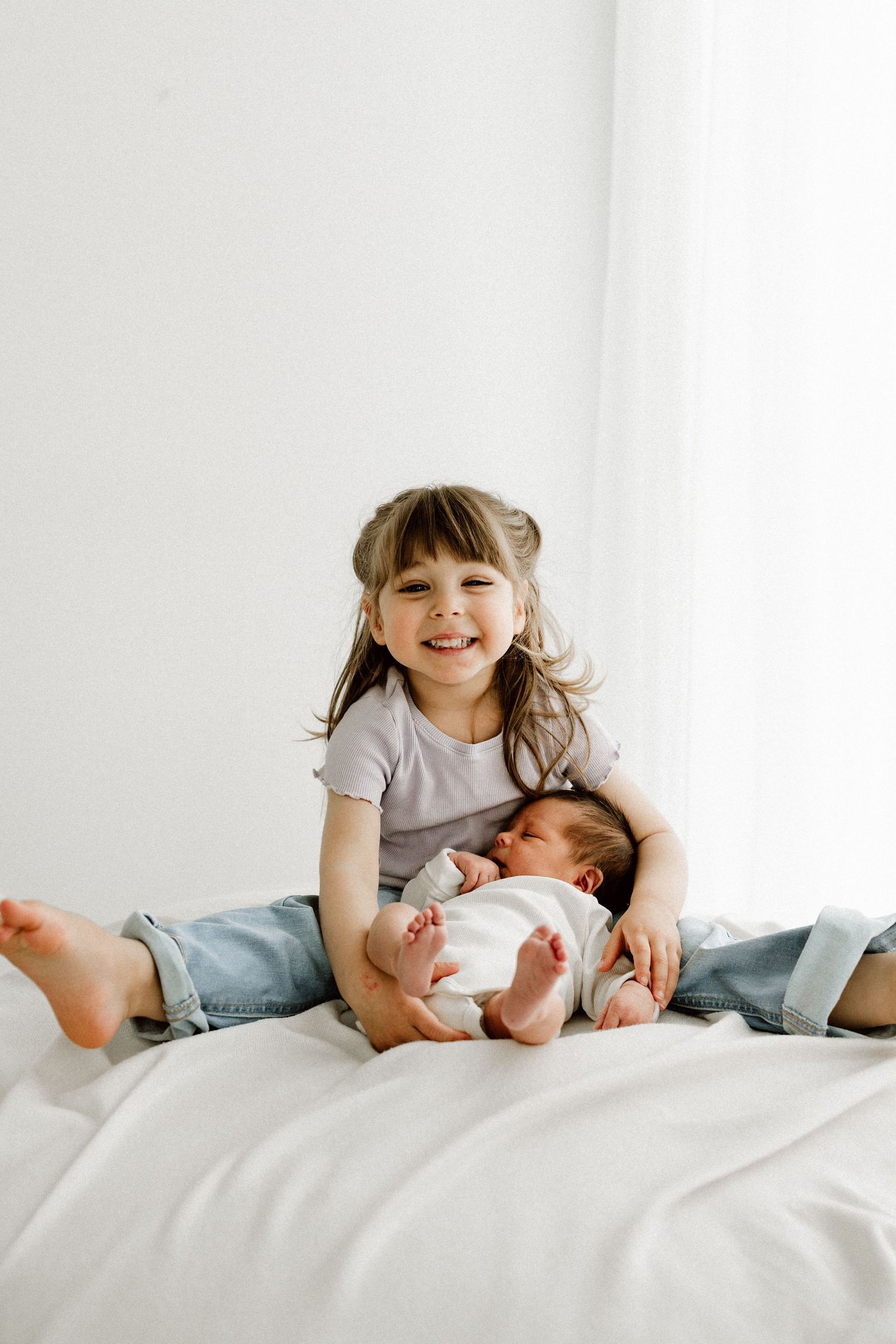 Smiling young girl sitting on white bed holding newborn baby sibling in her lap.