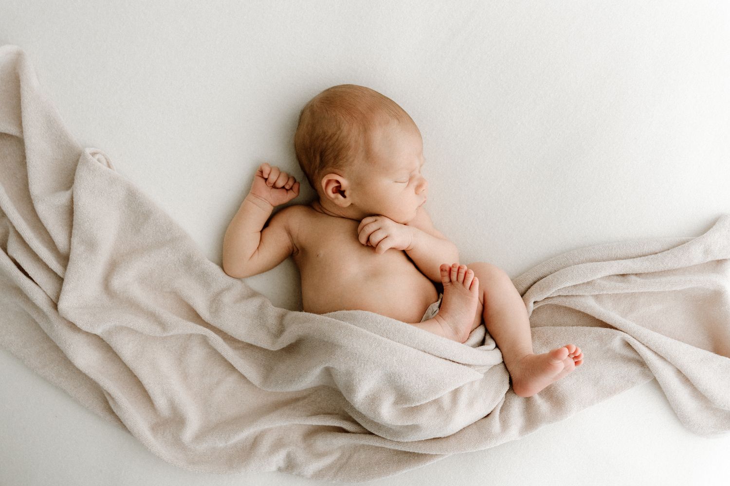 Sleeping naked newborn baby curled on white surface with soft beige blanket.