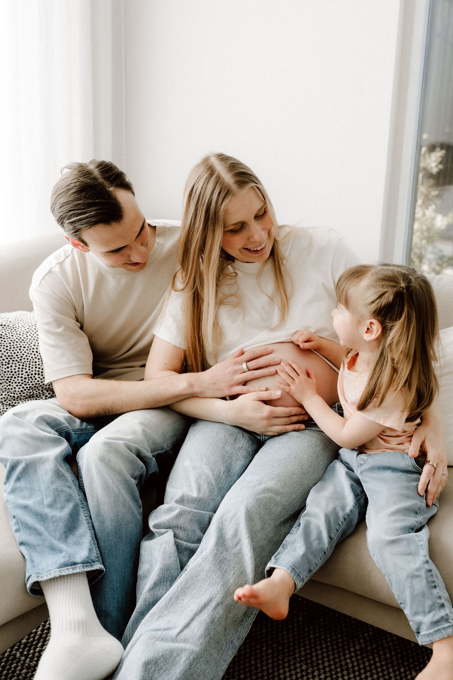 Family with father, pregnant mother, and young daughter sitting together touching baby bump on sofa.