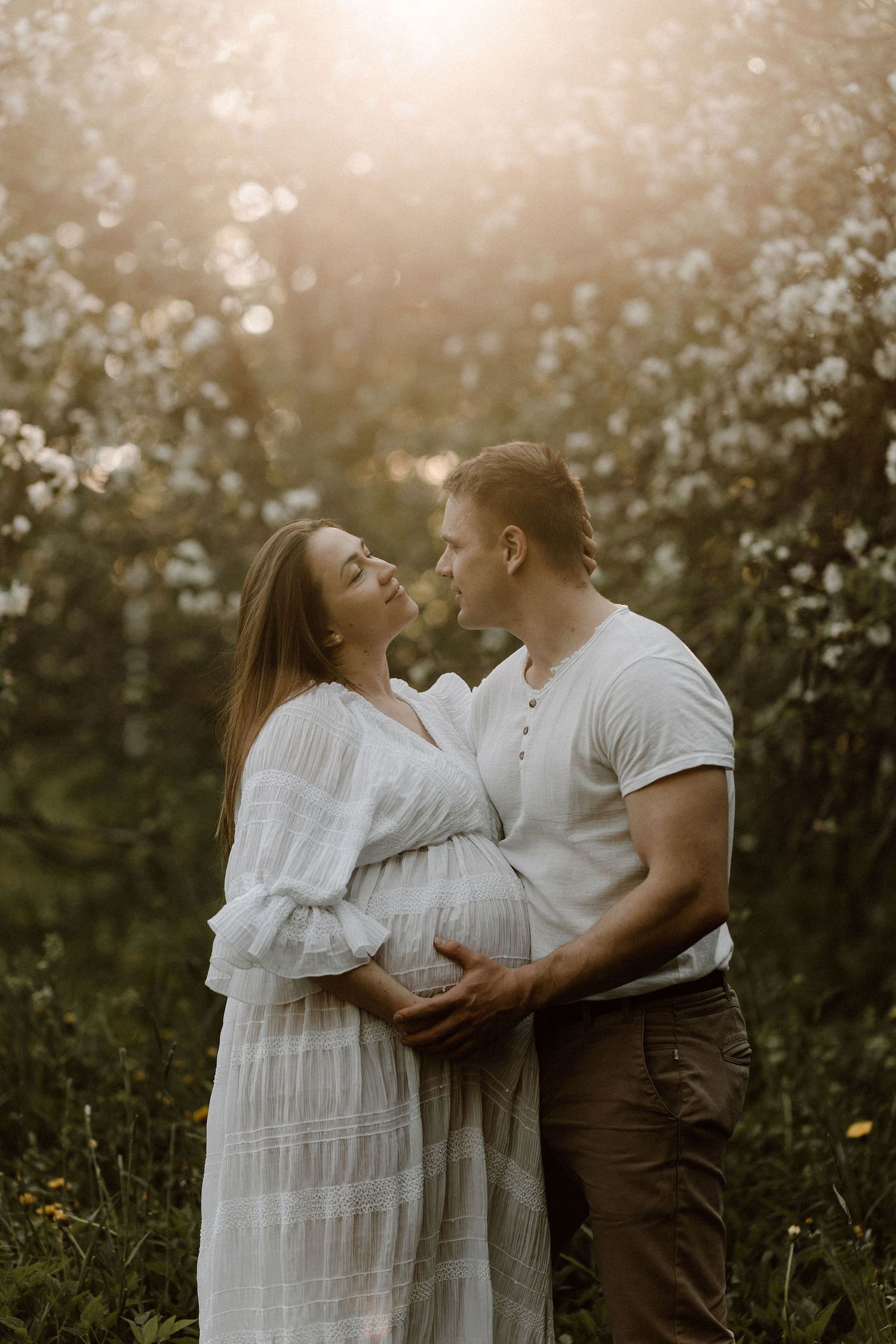 Couple sharing a tender kiss while man holds pregnant womans belly in sun-drenched flowering garden.