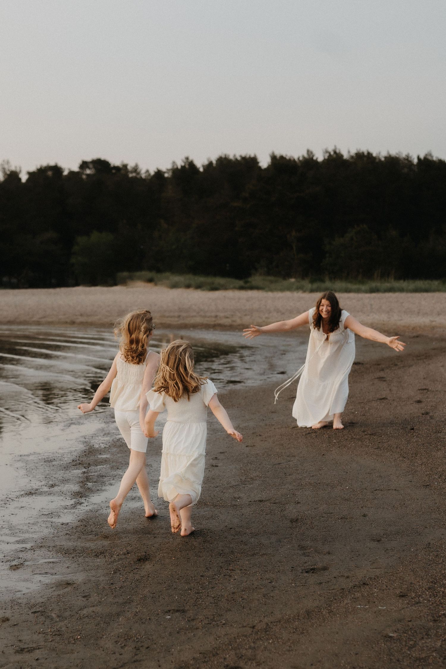 Two girls in white dresses run joyfully toward their mother along a sandy shoreline at dusk.