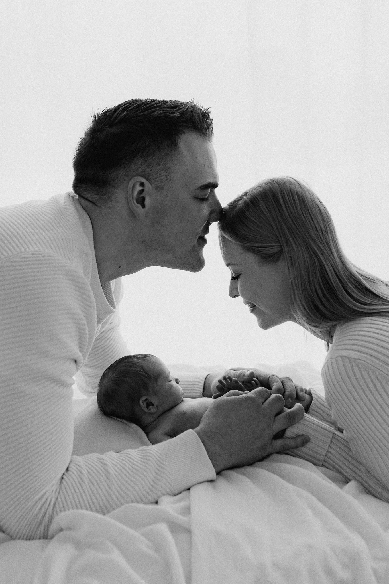 Black and white portrait of parents touching noses lovingly while cradling their newborn baby on a white blanket.