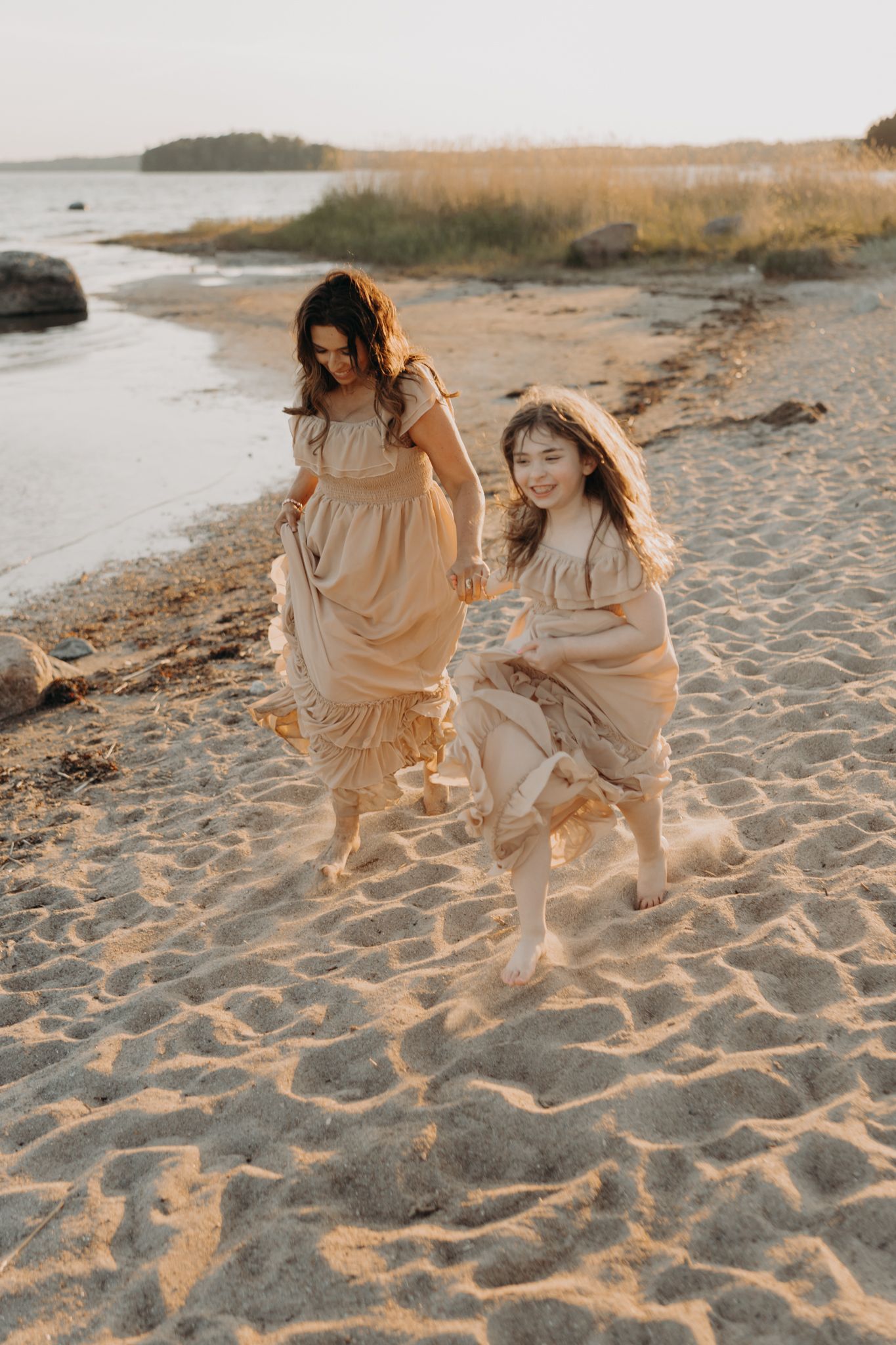 Mother and daughter in matching beige dresses running barefoot on sandy beach at sunset.