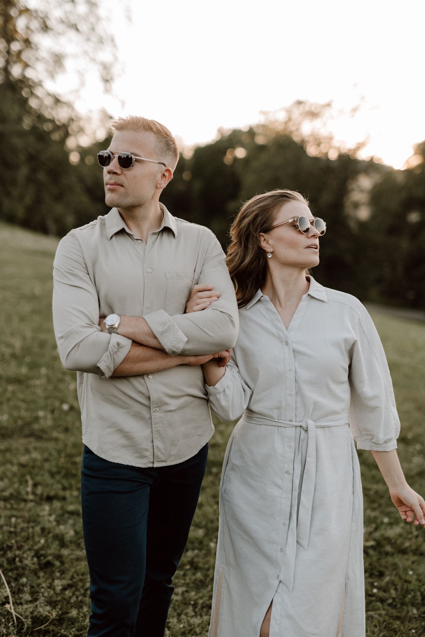 Couple in coordinating beige outfits wearing sunglasses standing apart in a sunlit green field.