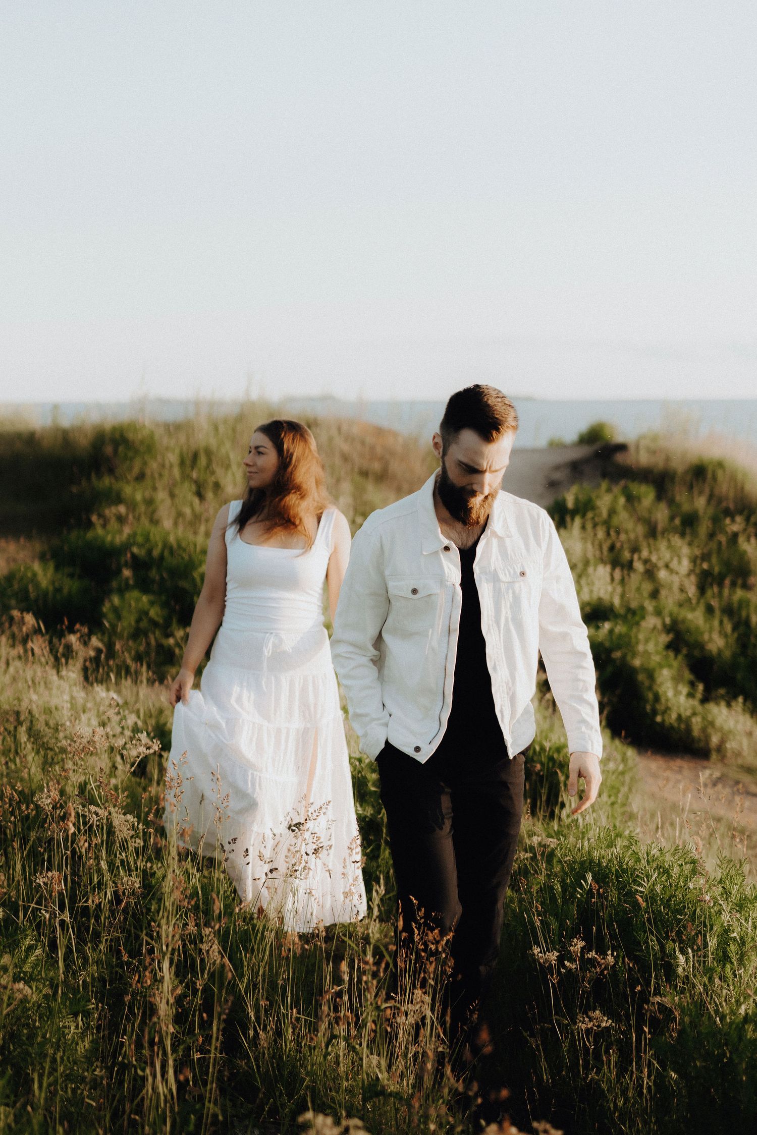Couple walking through sun-lit green meadow, woman in white dress, man in white jacket.