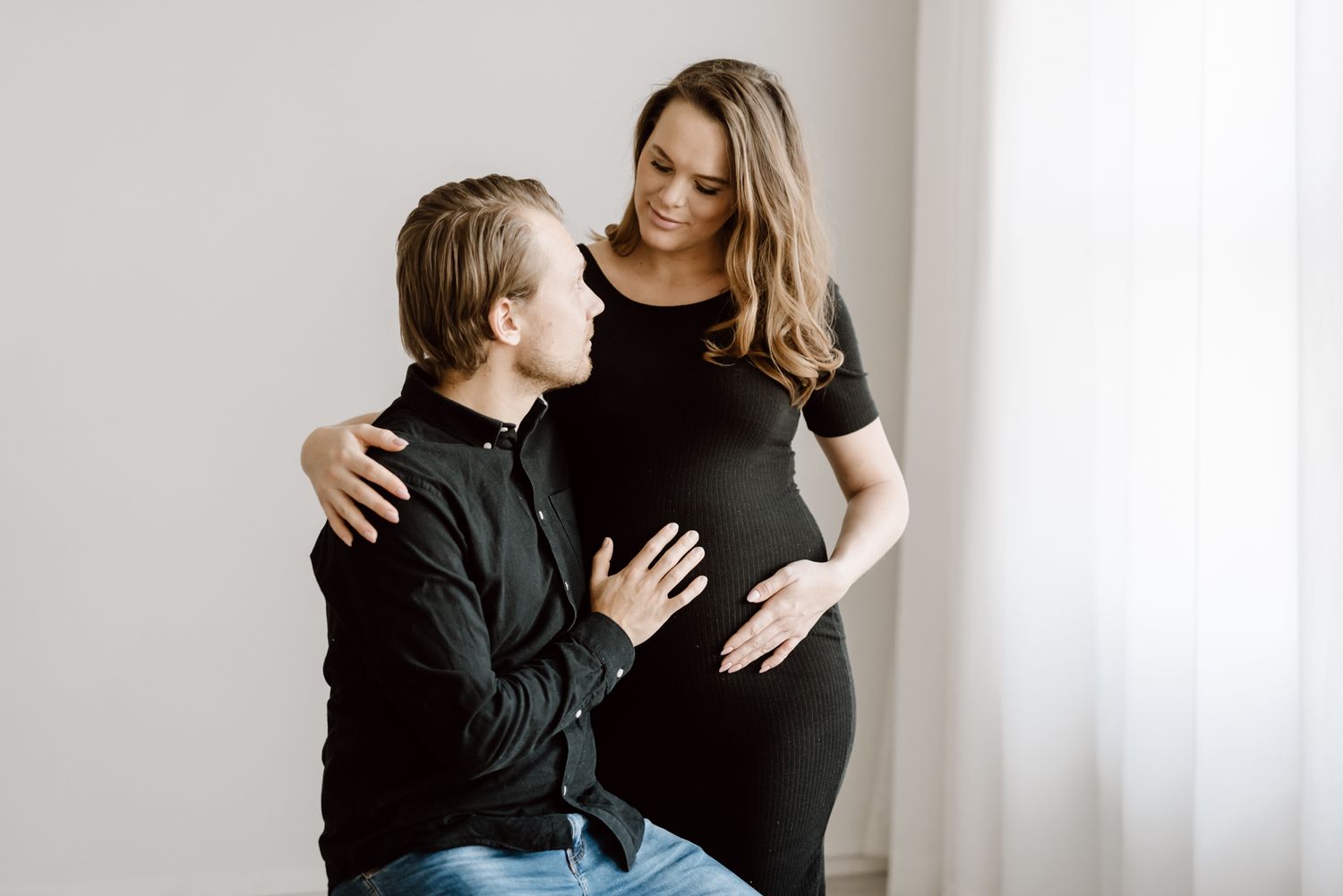 Couple in matching black outfits during maternity photo session by bright window indoors.