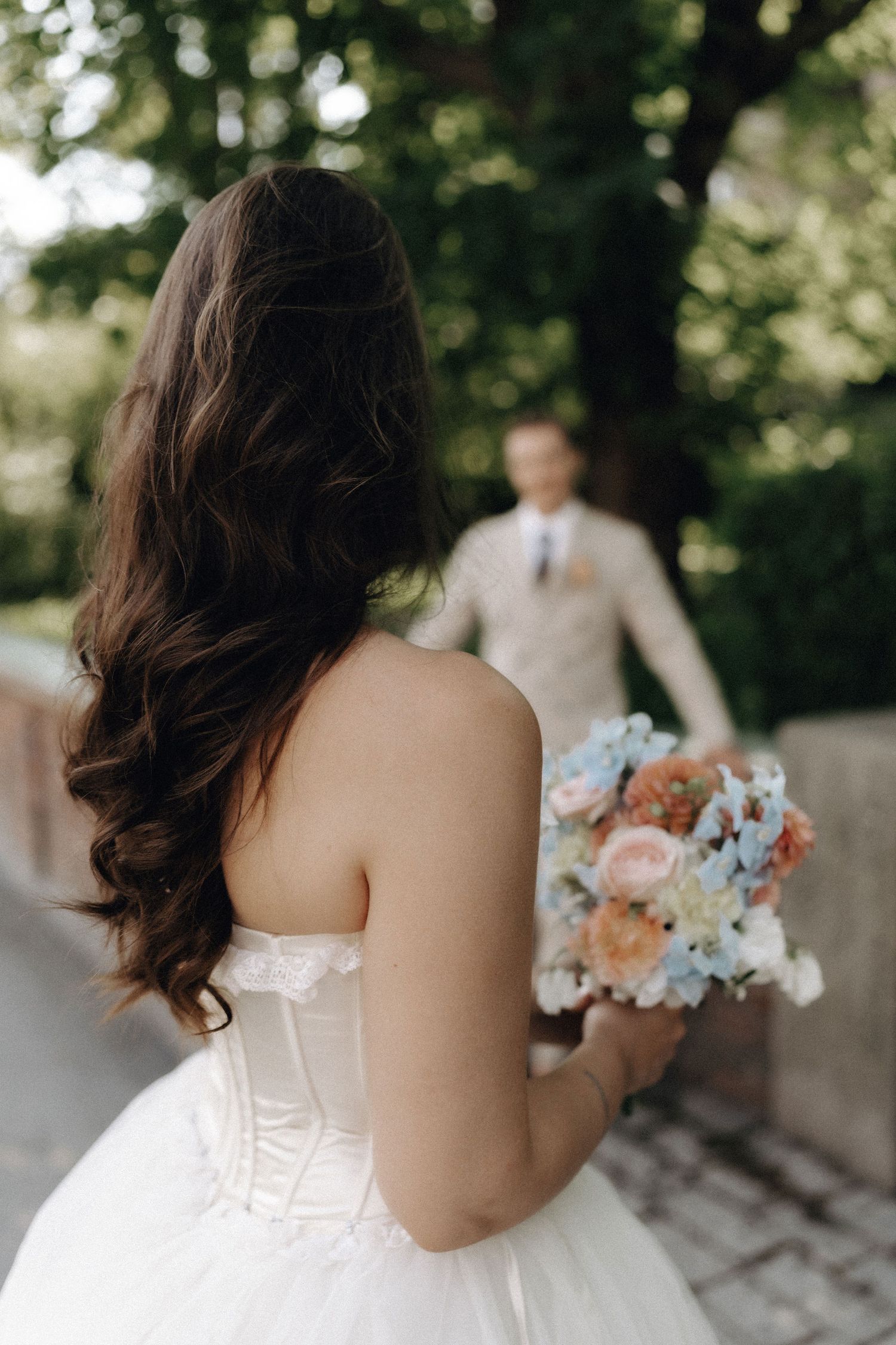 Bride with long brunette hair holds a pastel bouquet in a white tiered gown facing her groom outdoors.