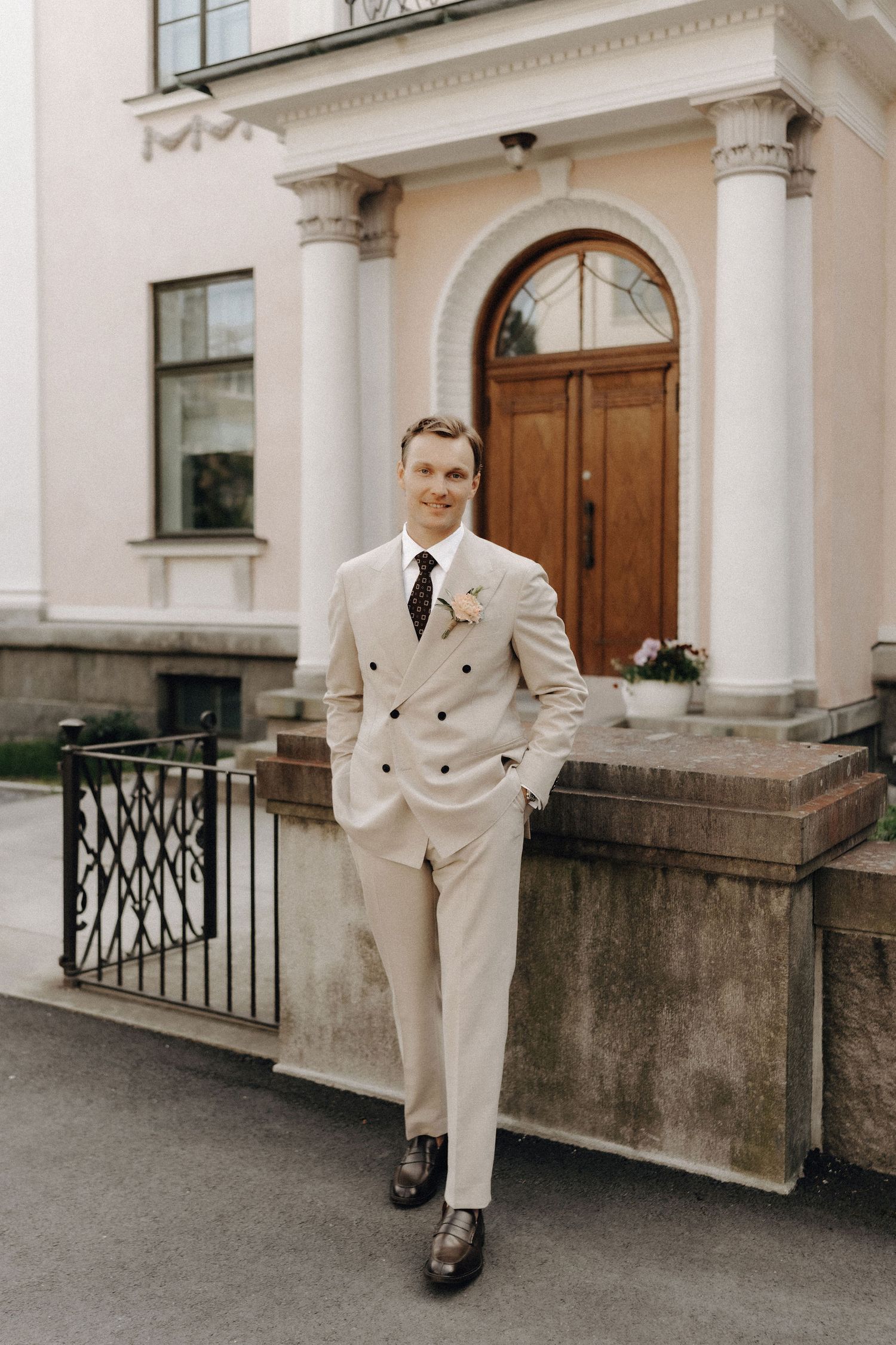 Stylish groom in a beige double-breasted suit with boutonniere posing in front of a neoclassical building.