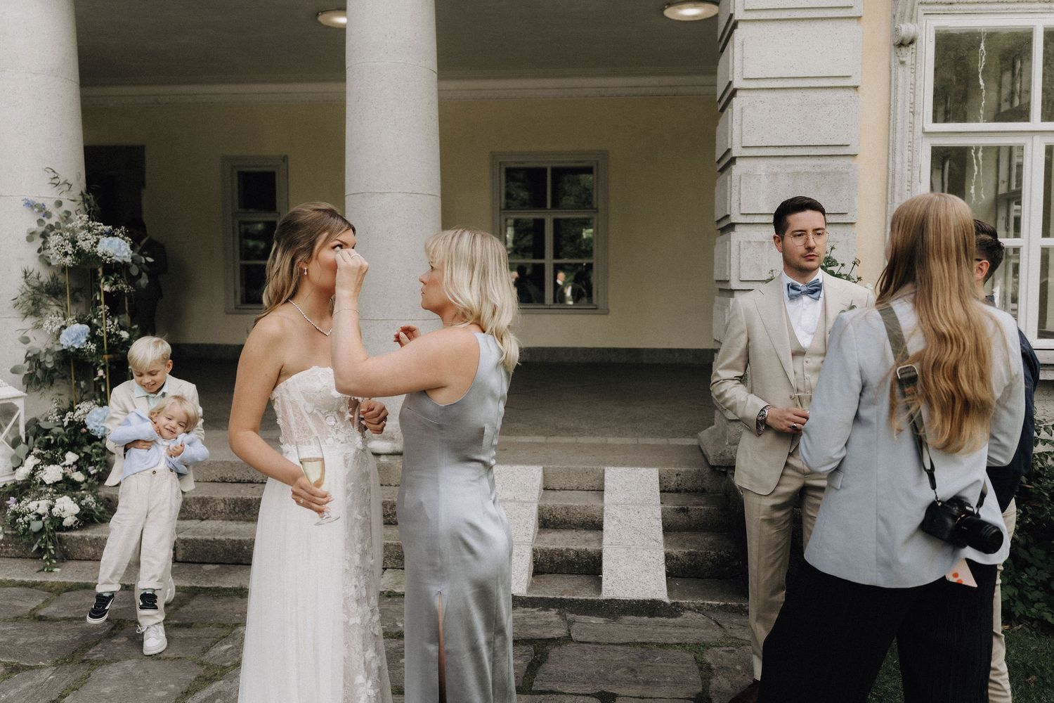 A woman tenderly touches the brides face during an outdoor wedding reception near stone columns.