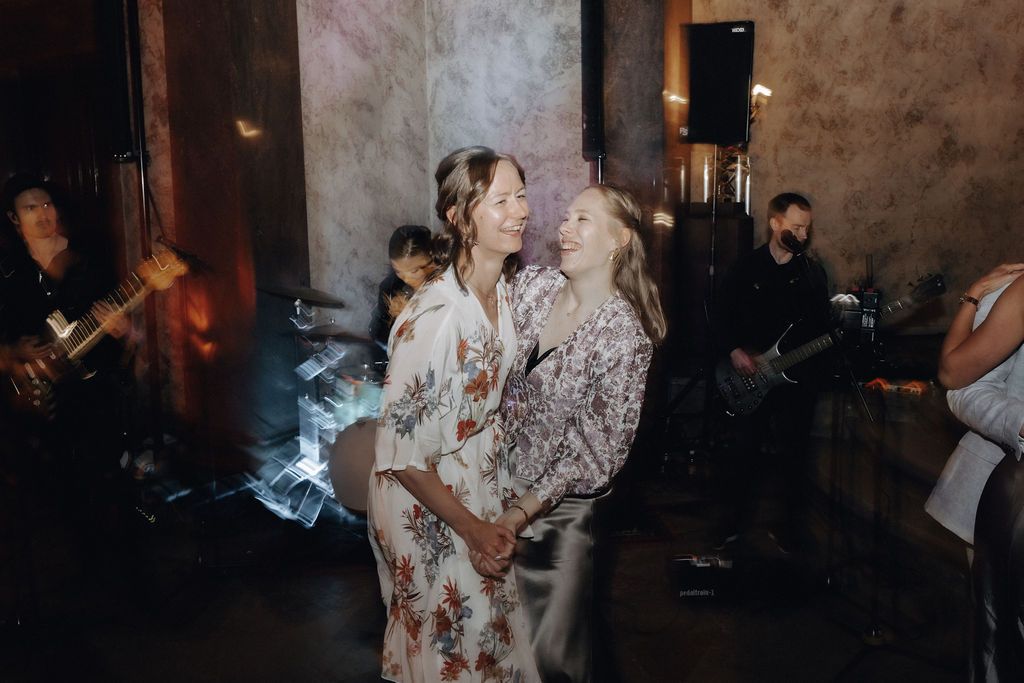 Two women in floral dresses share a joyful moment together near live band at wedding.