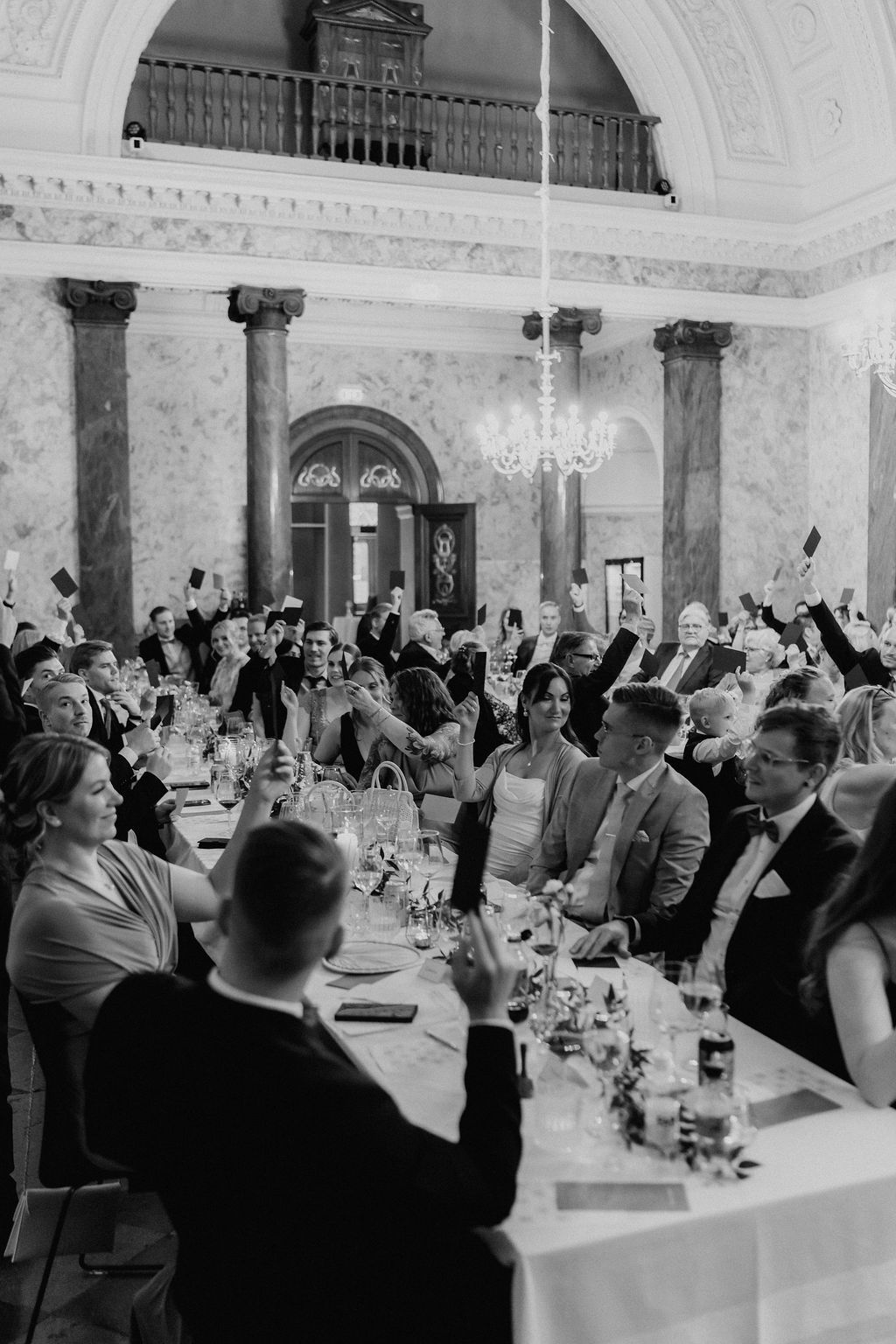 Wedding guests raise hands in a grand marble hall during a lively reception toast.