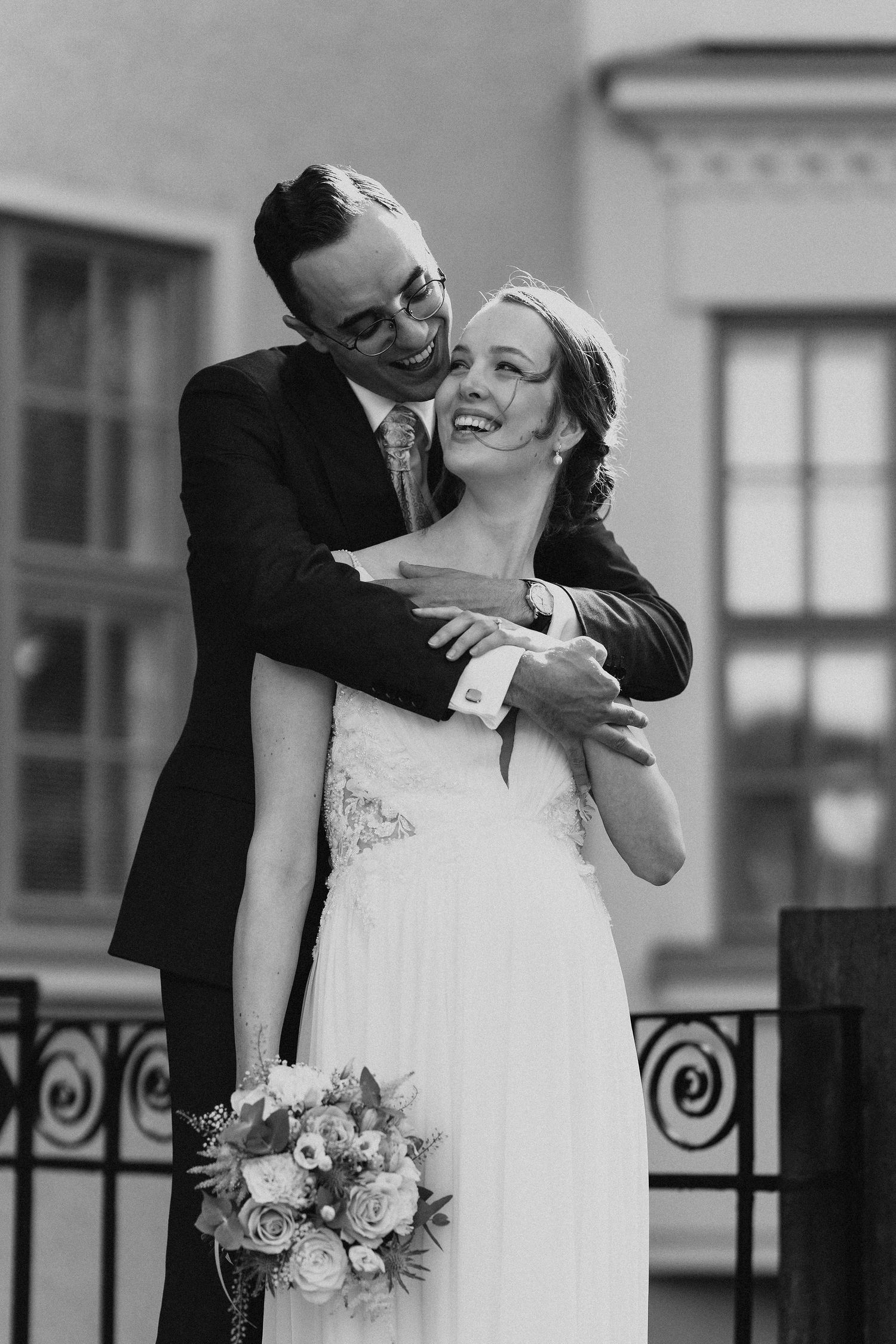 Black and white photo of groom hugging laughing bride from behind near an ornate iron fence.