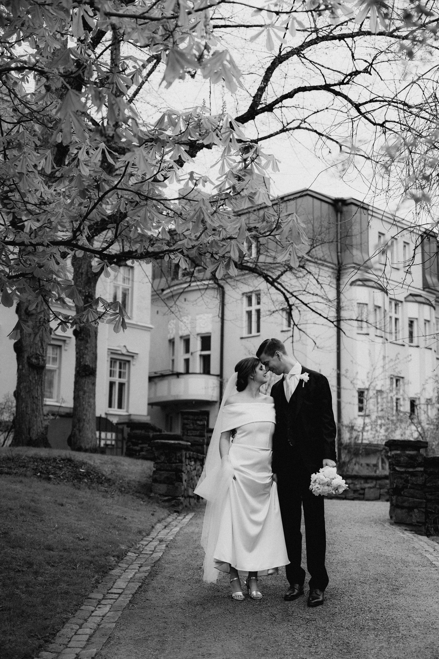 Black and white wedding photo of bride and groom kissing under a tree outside a grand building.