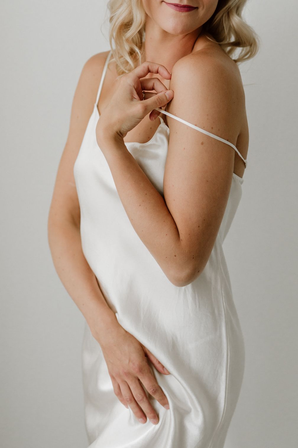 Woman in white satin slip dress adjusting shoulder strap against a light background.